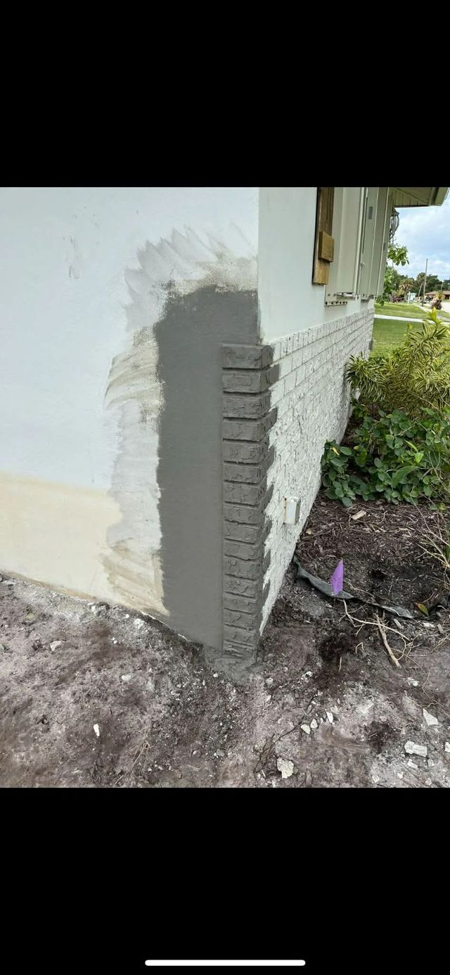 Close-up of a house corner with a white wall, exposed brick edge, and ground beside a garden path.