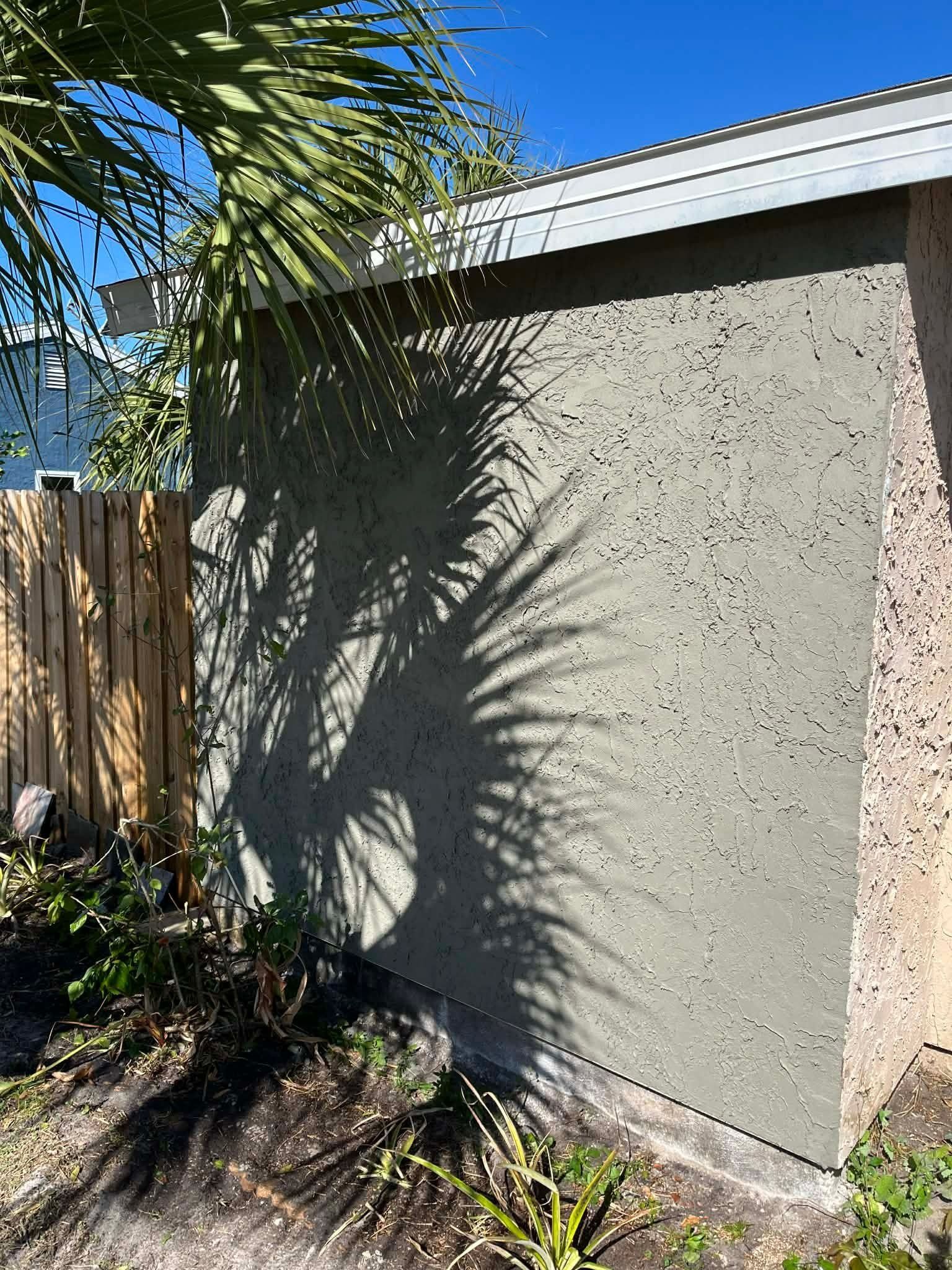 Stucco exterior wall with roof edge, palm frond shadows, and a wooden fence in a sunny yard