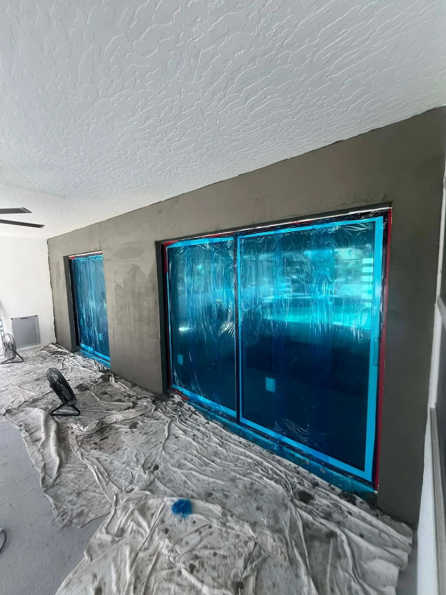 Interior room under renovation with blue-taped windows and plastic sheeting on the floor.