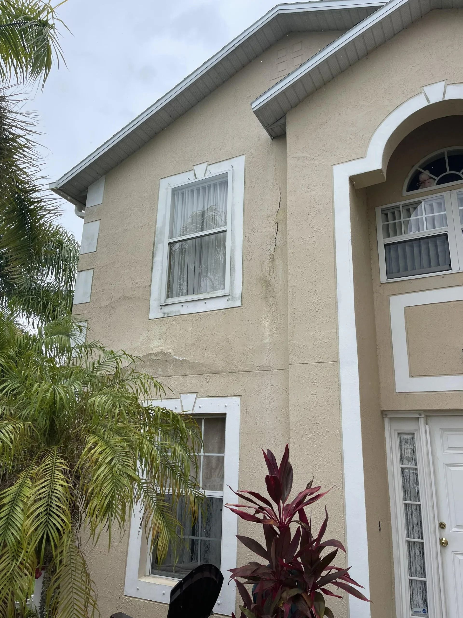 Two-story beige stucco house with white trim, arched entry, and tropical plants in front