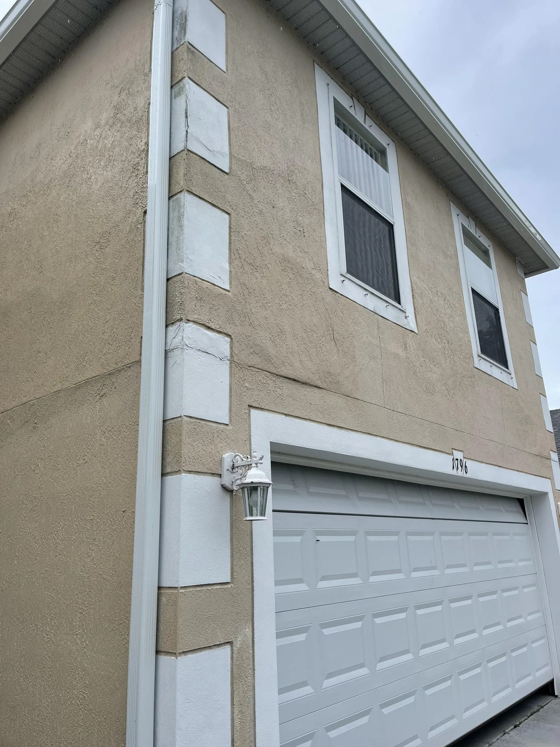 Corner of a beige house with white trim and a white garage door under an overcast sky