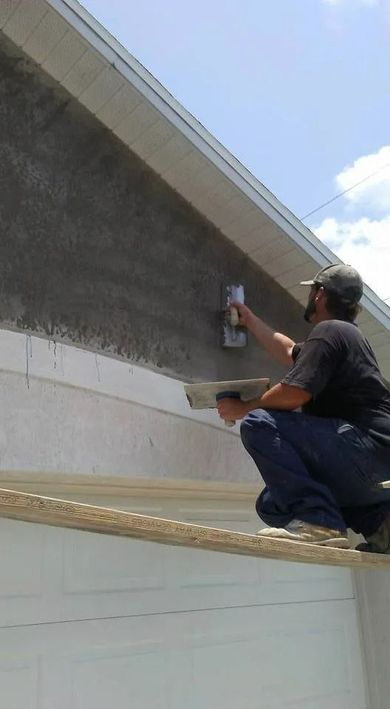 Man painting a house exterior wall on a ladder, spraying or rolling gray paint near the roofline.