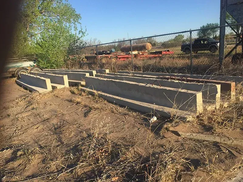 Concrete drainage culverts beside a dirt road near a chain-link fence in a rural area.
