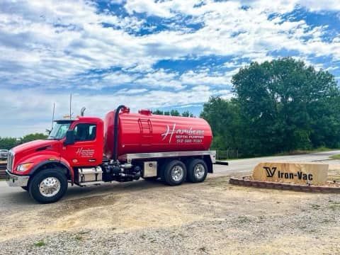 Red vacuum tanker truck parked by a dirt road beside an Iron-Vac sign under a cloudy sky