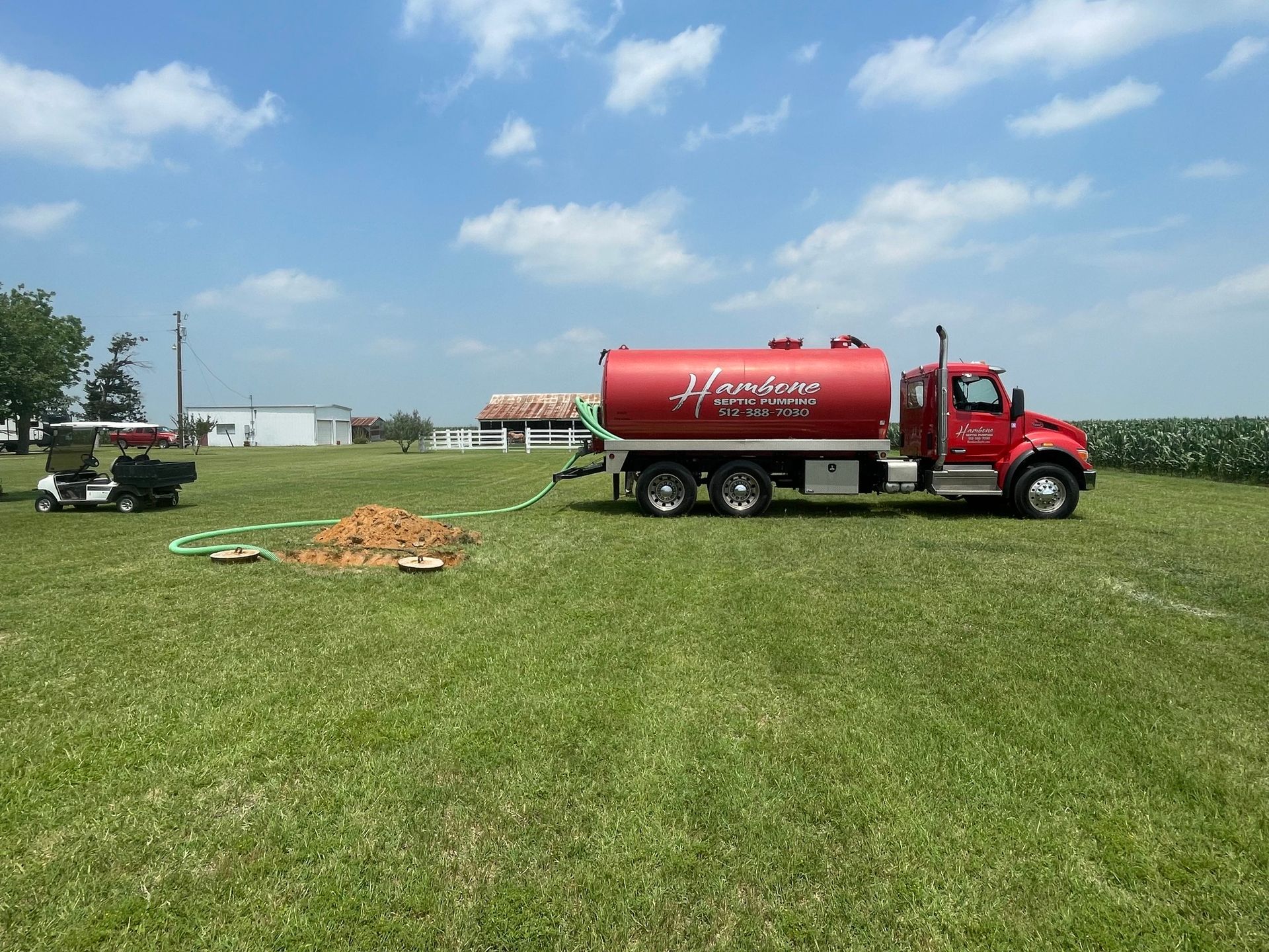 Red tanker truck spraying liquid on a grassy field near a farm under a blue sky.