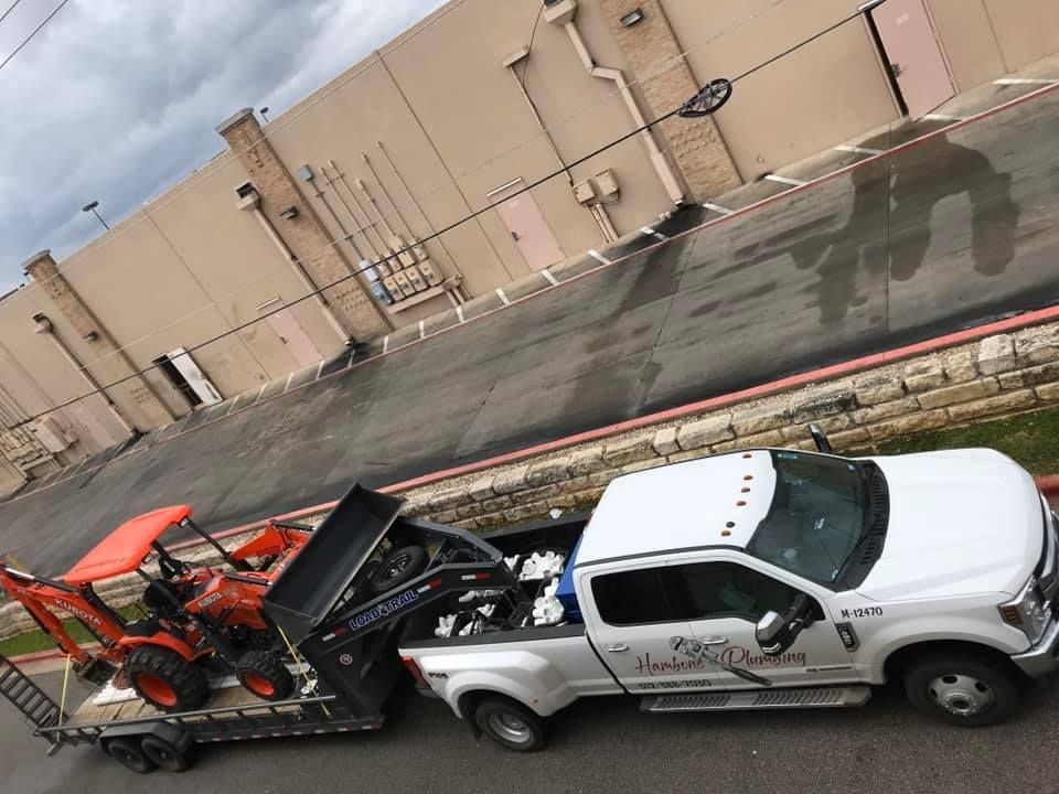 White tow truck hauling orange farm equipment beside a beige warehouse.