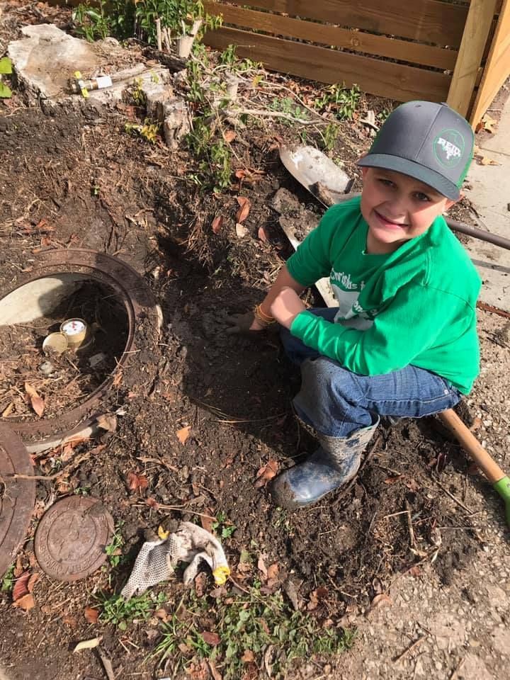 Child in a green shirt and cap smiling while kneeling in a muddy garden beside a small pipe opening.
