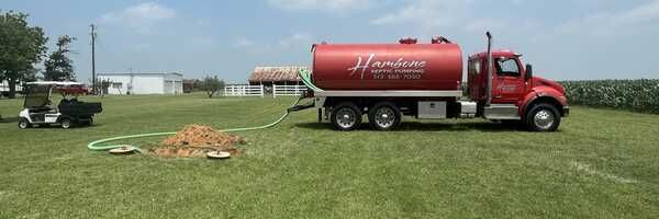 Red vacuum tanker truck on grass near a hose, with a cornfield in the background.