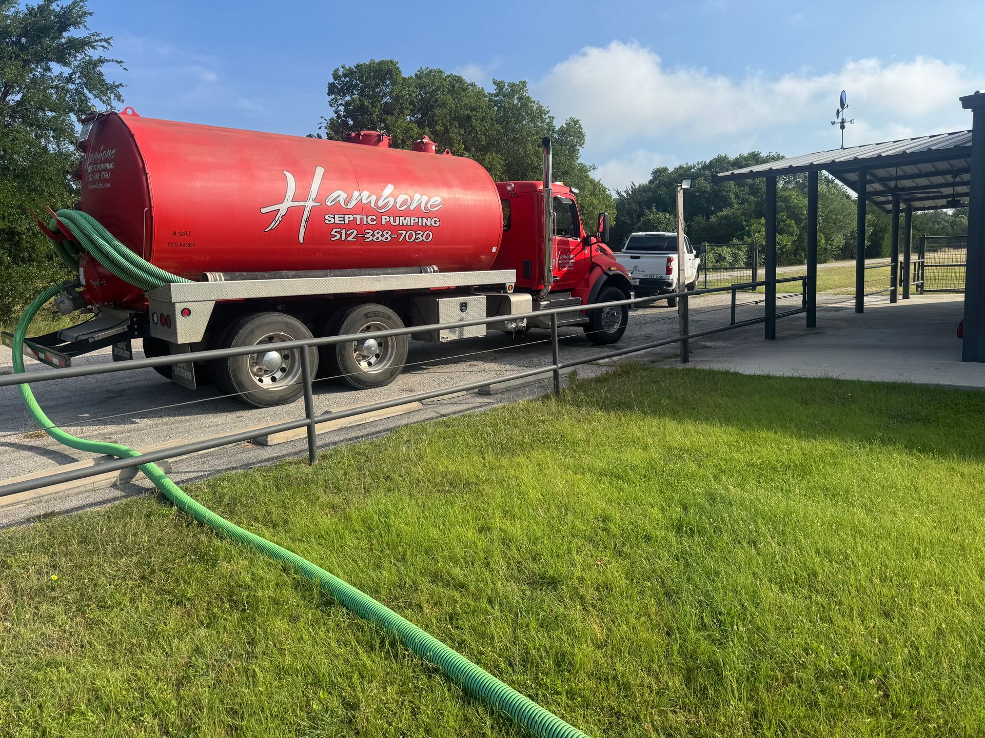 Red liquid tanker truck parked beside a metal canopy on a grassy lot.