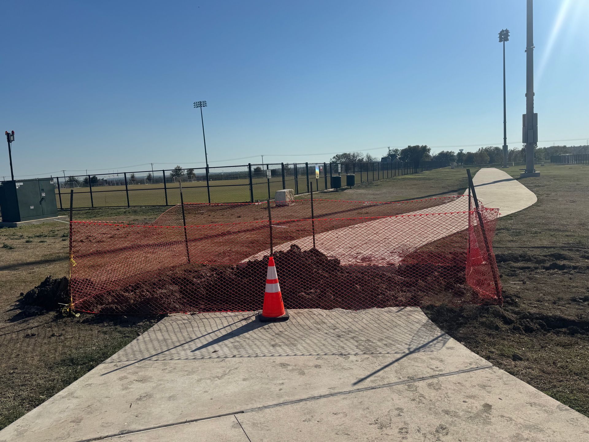 Orange traffic cone blocking a cracked sidewalk beside red mesh fencing on a sports field path