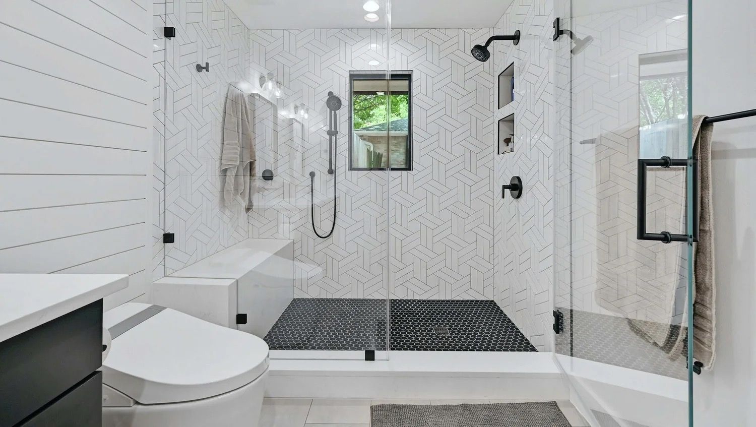 Modern white bathroom with a glass walk-in shower, bench, black fixtures, and a window