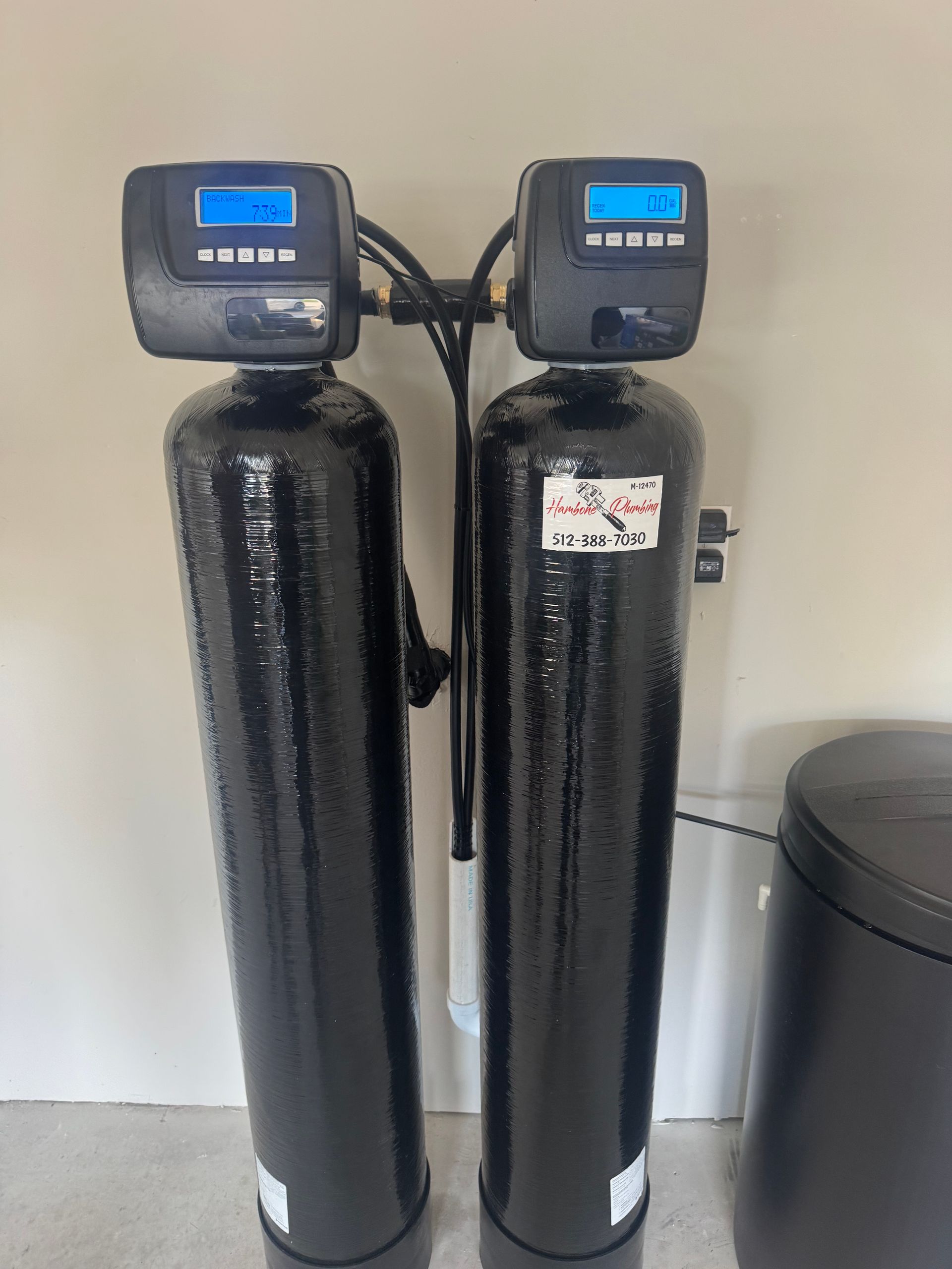 Two black water softener tanks with blue digital control heads, standing indoors beside a gray container.