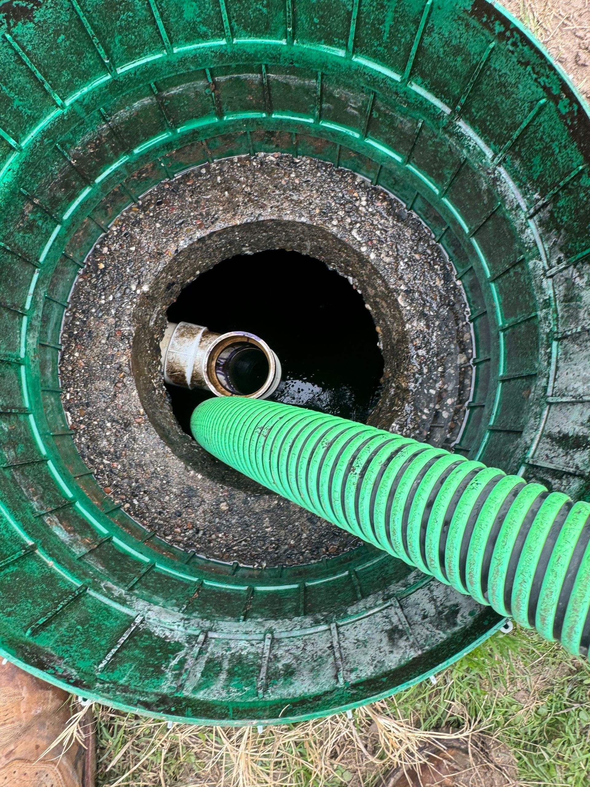 Green hose coiled into a circular drain or sump opening outdoors, viewed from above