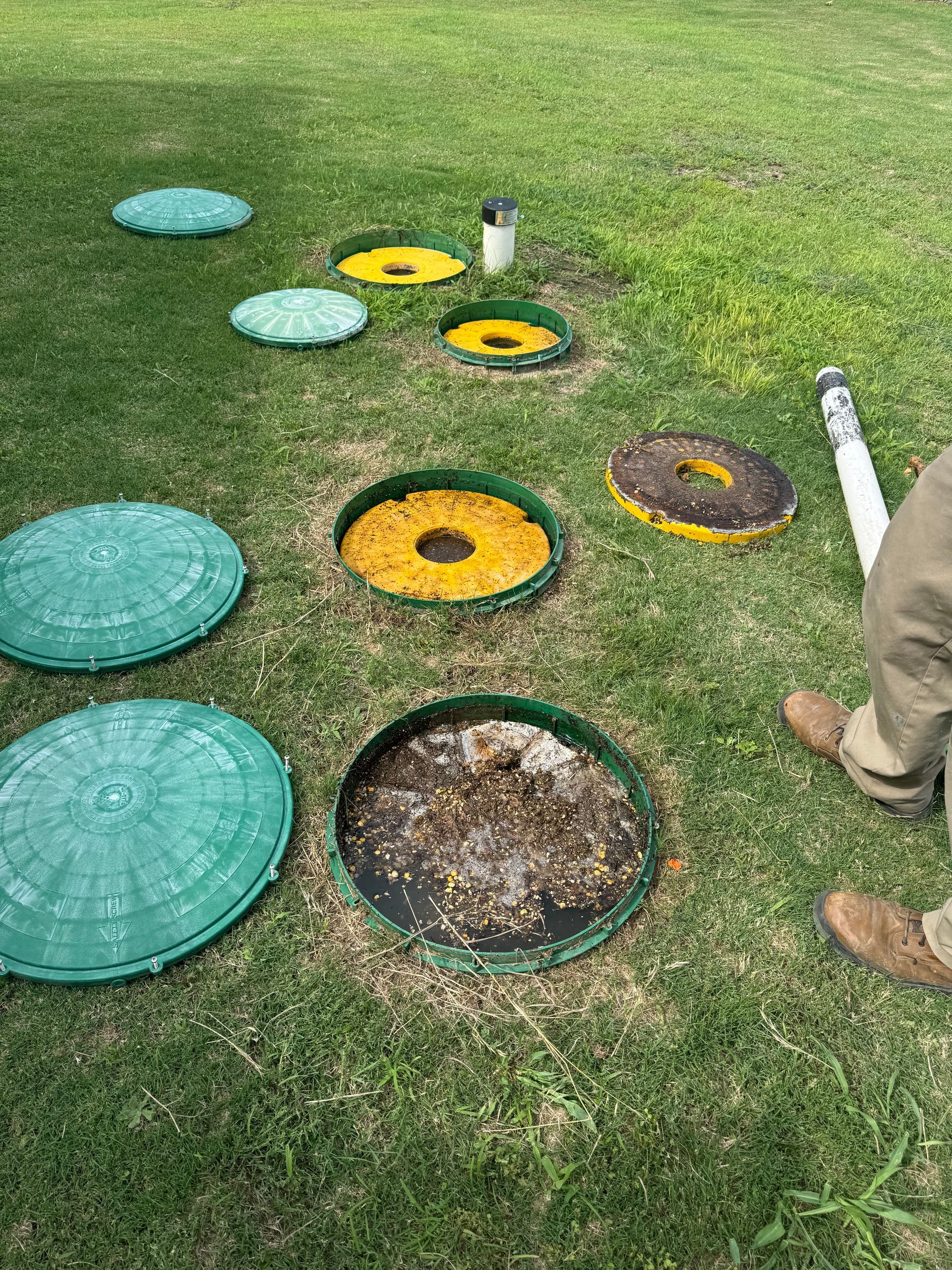 Raised circular lawn covers in a grassy field, some teal and yellow, with a person’s legs at right.