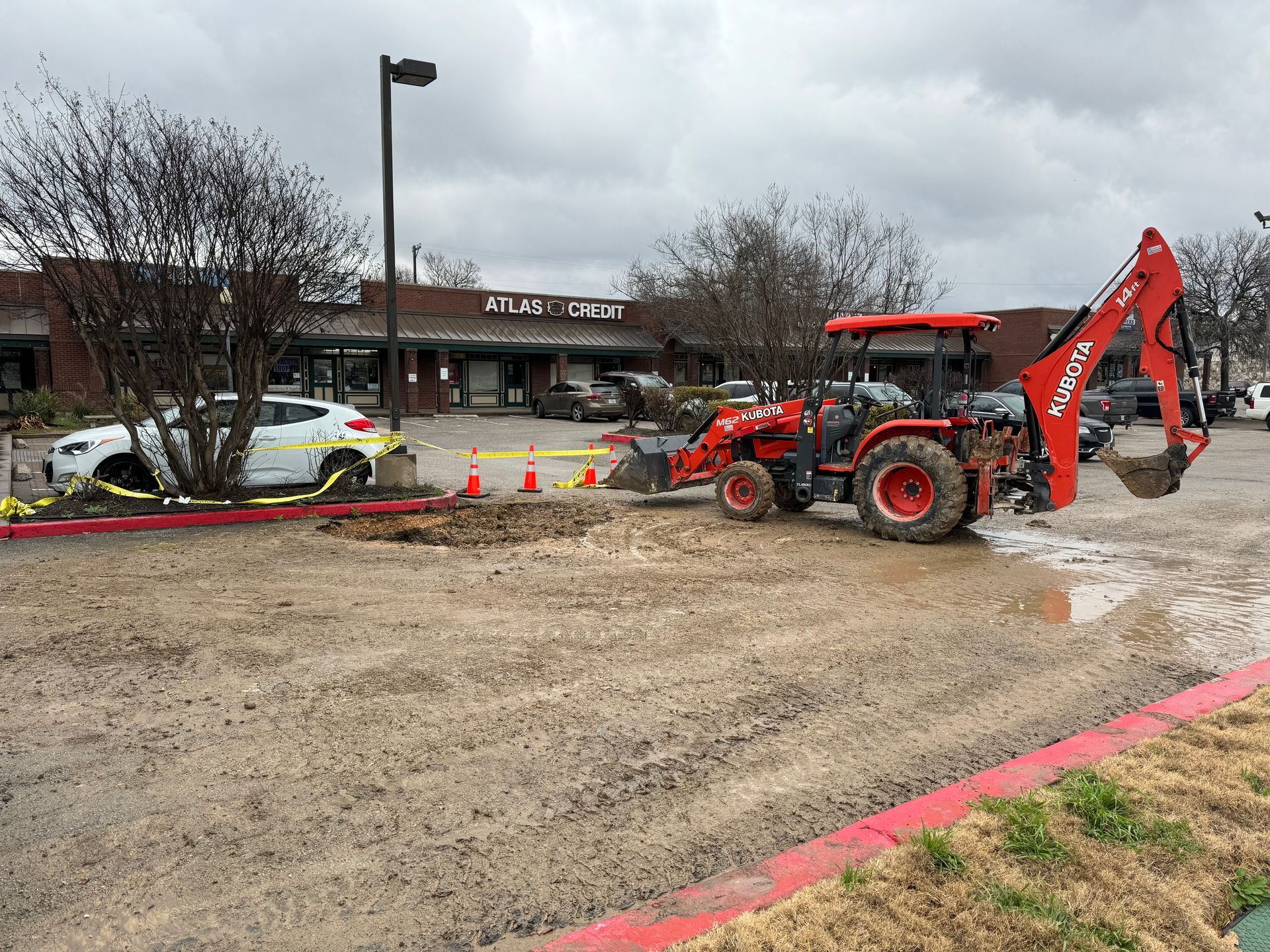 Red backhoe at a muddy construction site beside a building and parked cars, with orange cones and caution tape.