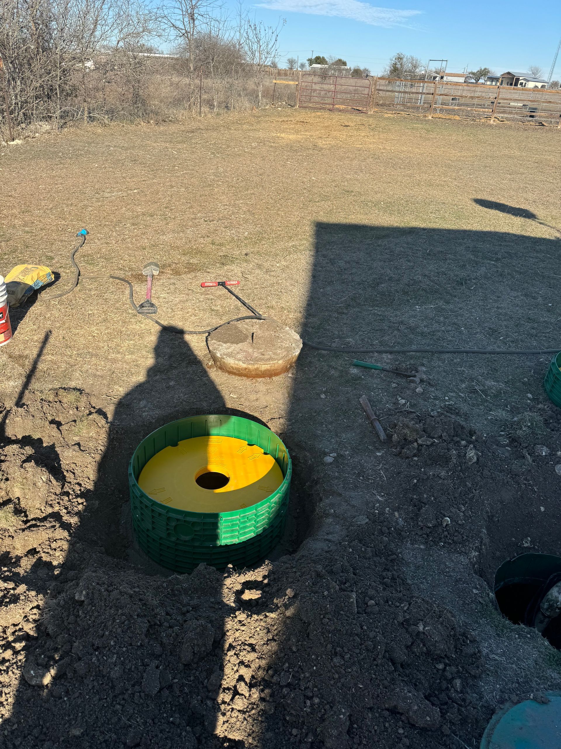 Mini golf hole with a yellow cup and green rim on gravel, with long shadows in a sunny outdoor area