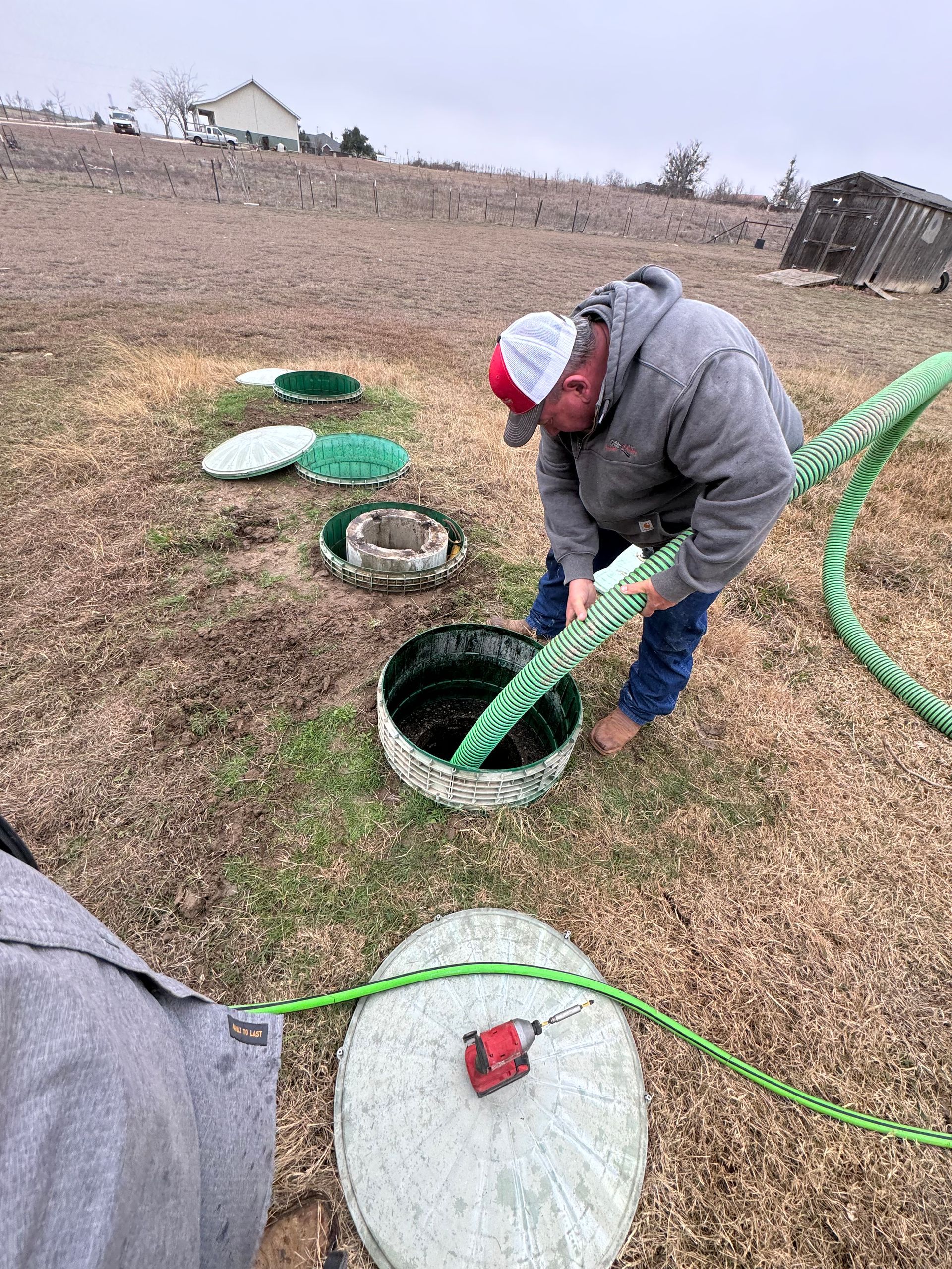Person in gray jacket and red hat handling a green hose beside open utility covers in a field.