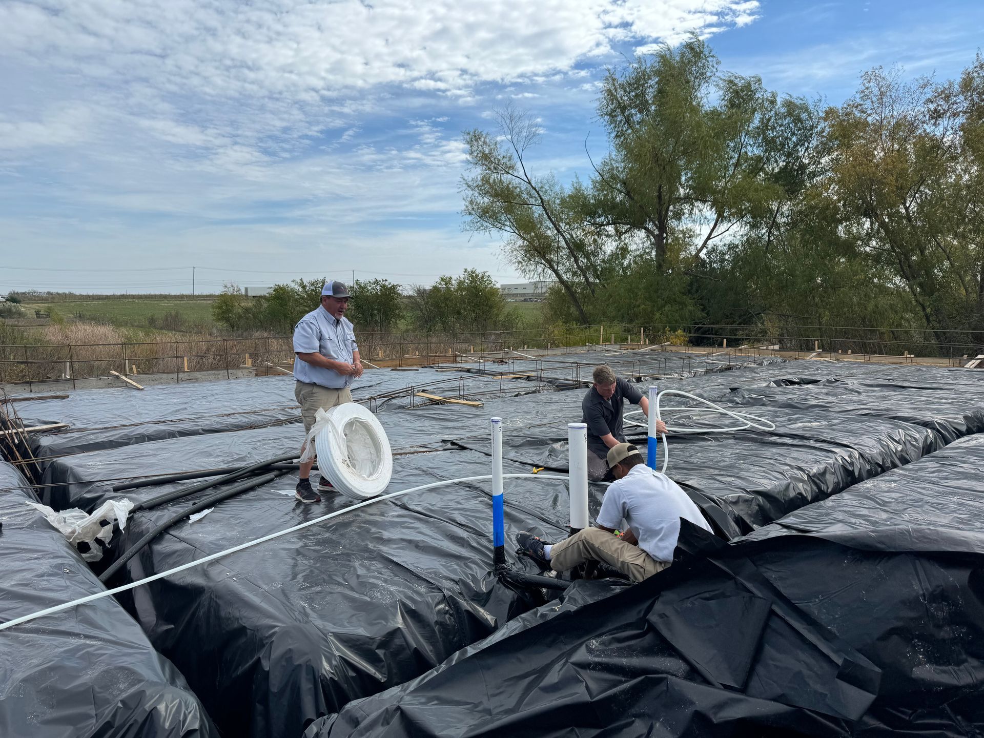 Workers inspecting black plastic-covered soil in a field with hoses under a cloudy sky