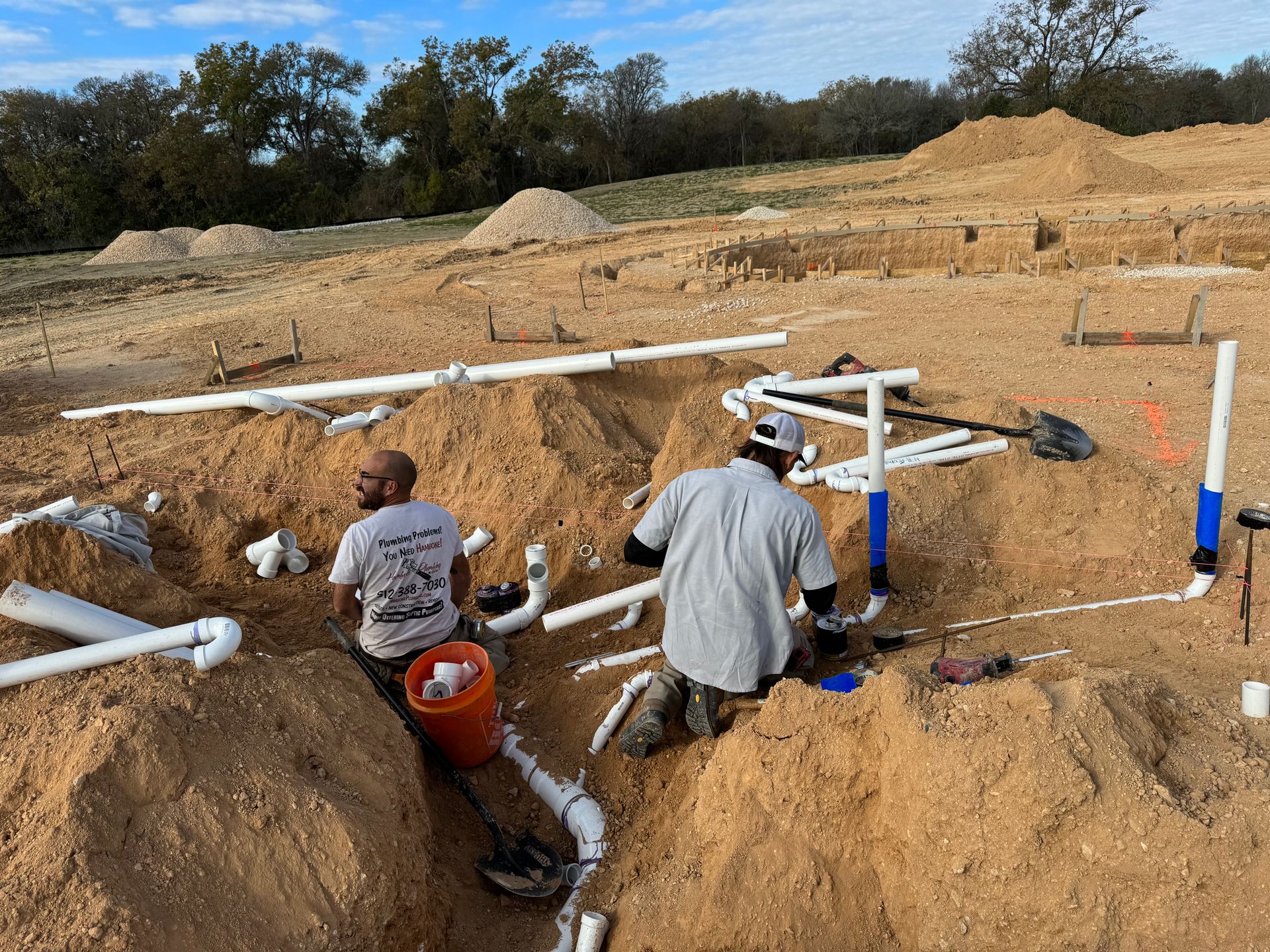 Workers install white pipes in a sandy excavation site with blue and red markers.
