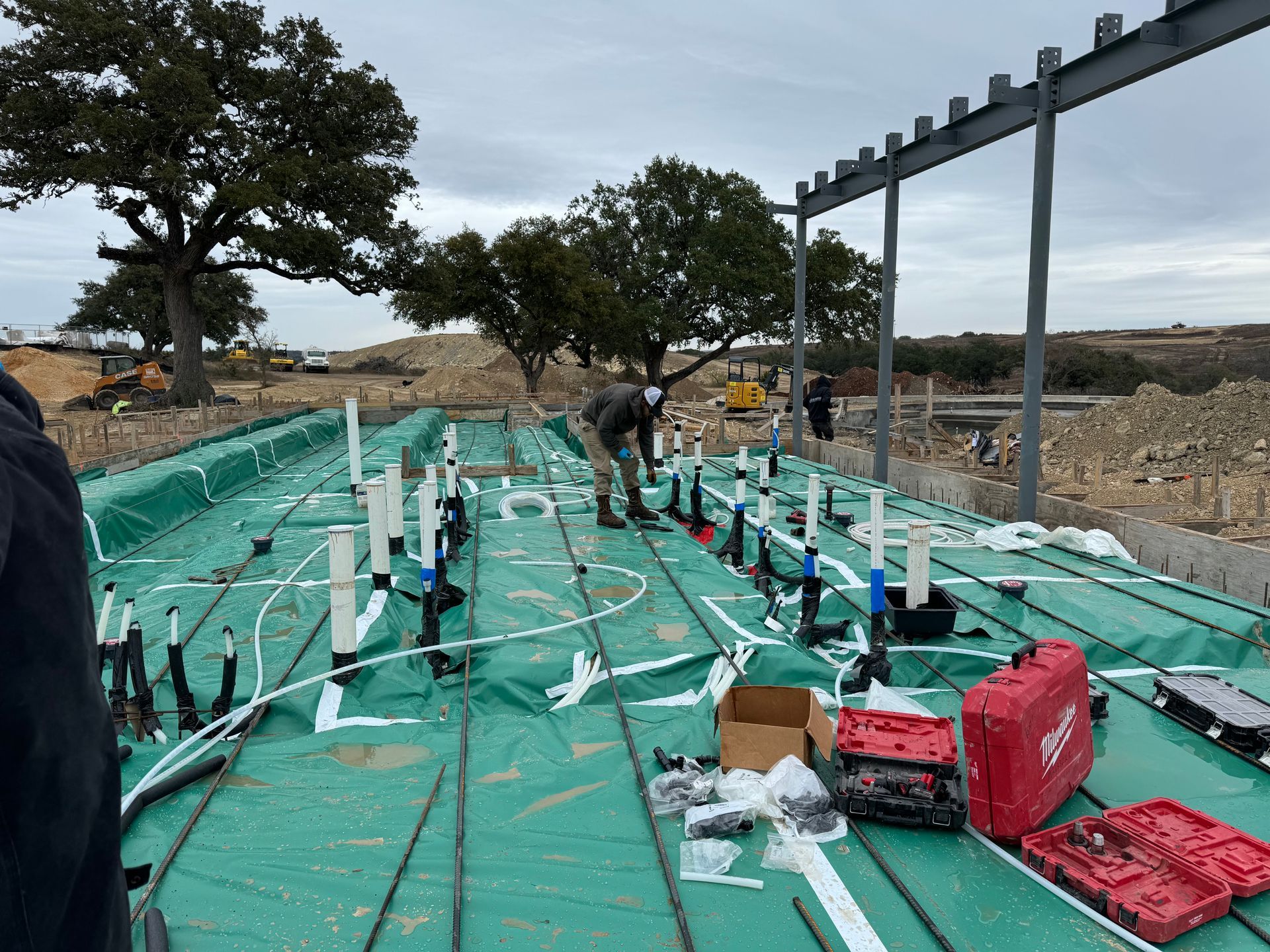 Construction site with green tarp-covered ground, pipes, workers, and steel frame under overcast sky