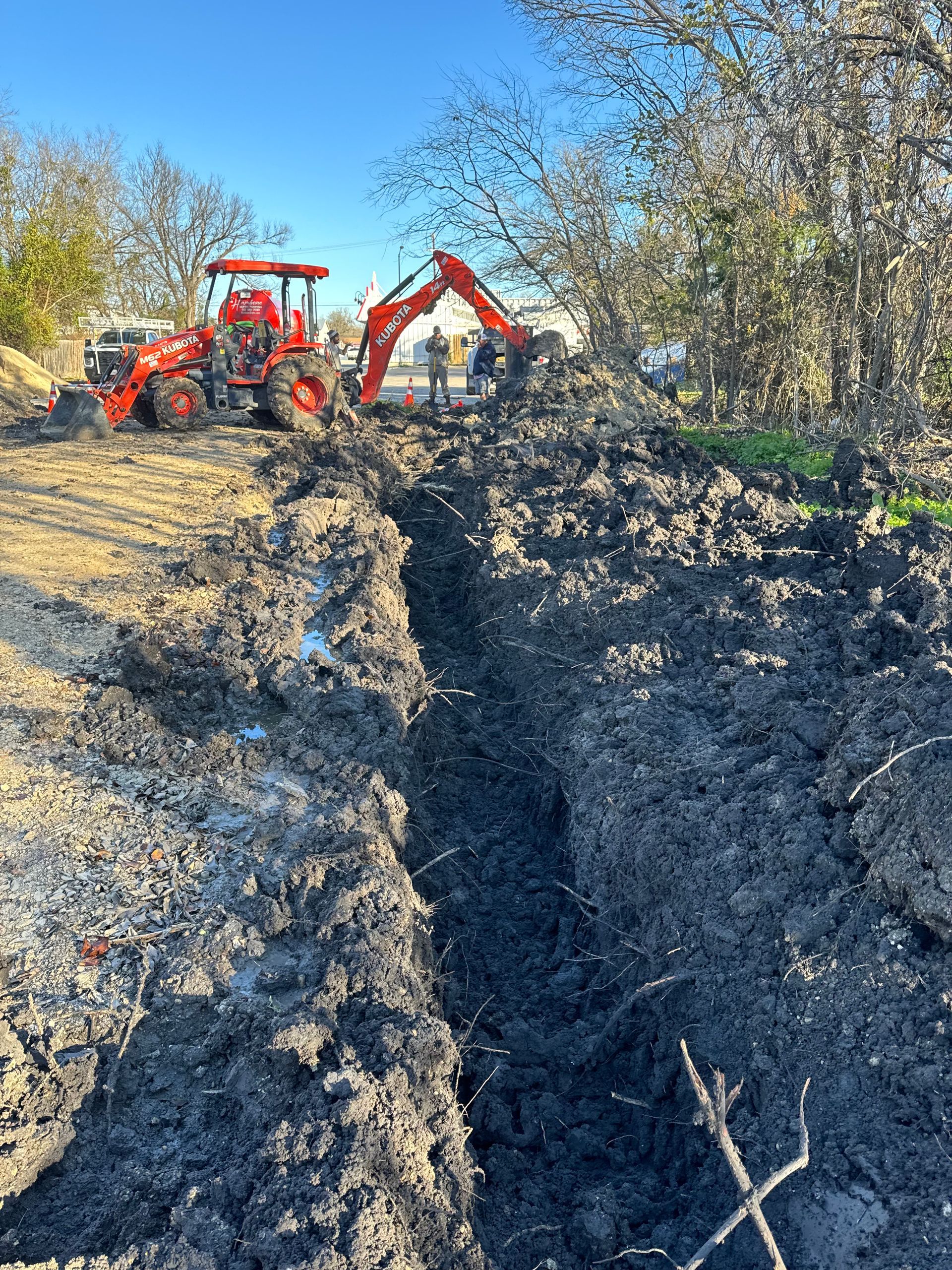 Orange excavators digging a deep trench in a wooded area under a clear blue sky