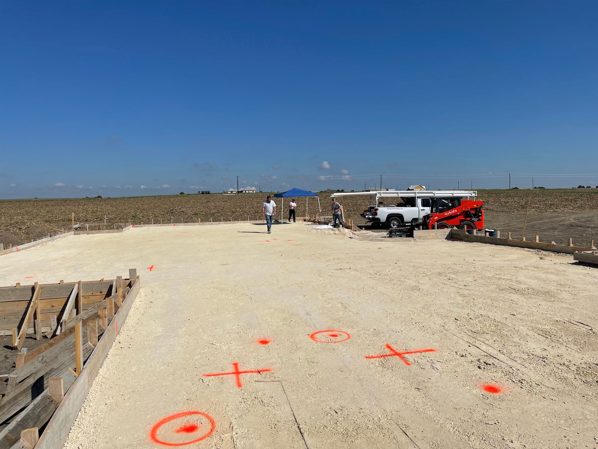 Road under construction with fresh concrete, orange markings, workers, and pickup trucks under a clear blue sky
