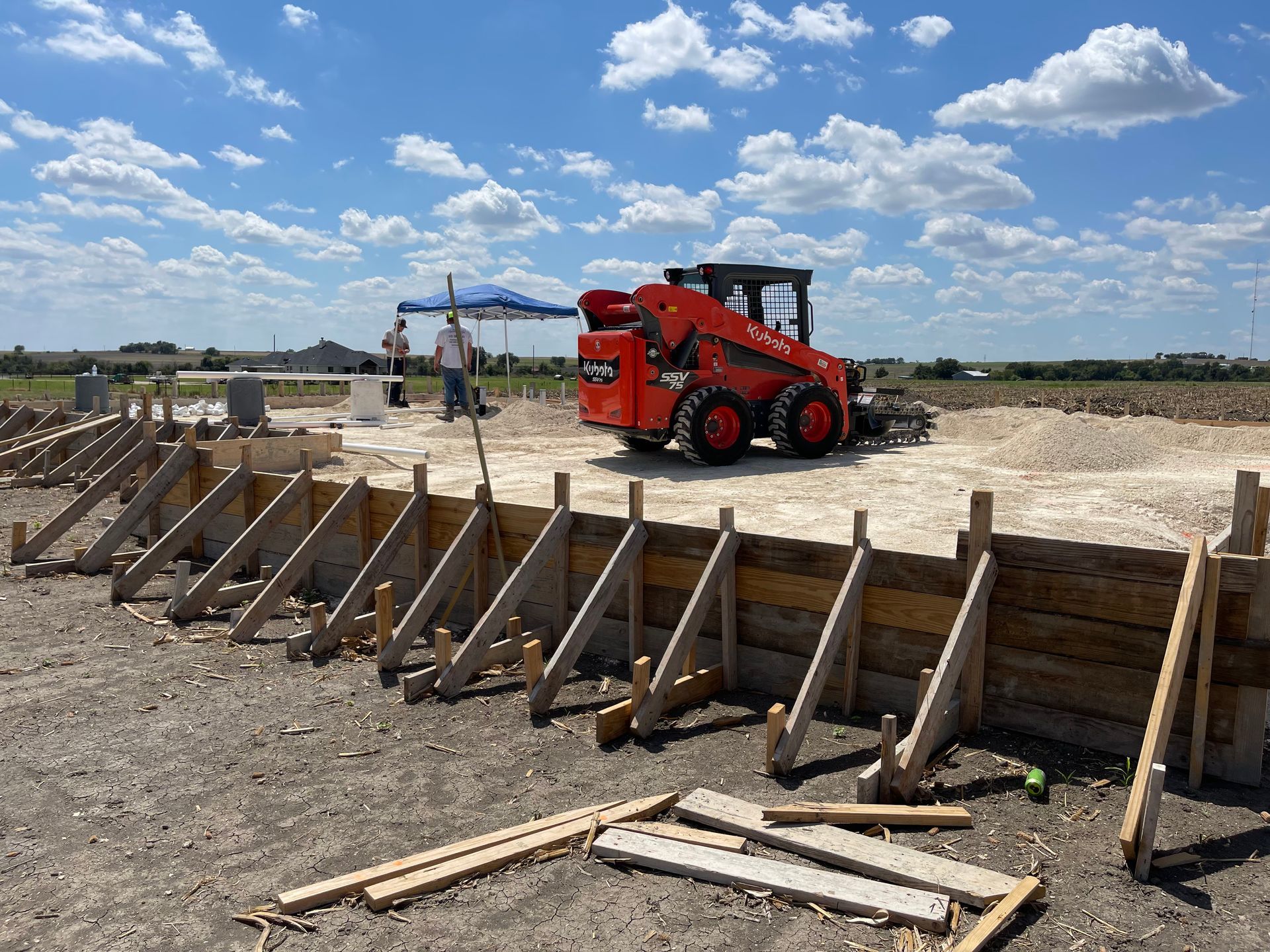 Construction site with red skid steer beside a wooden trench wall under a blue sky