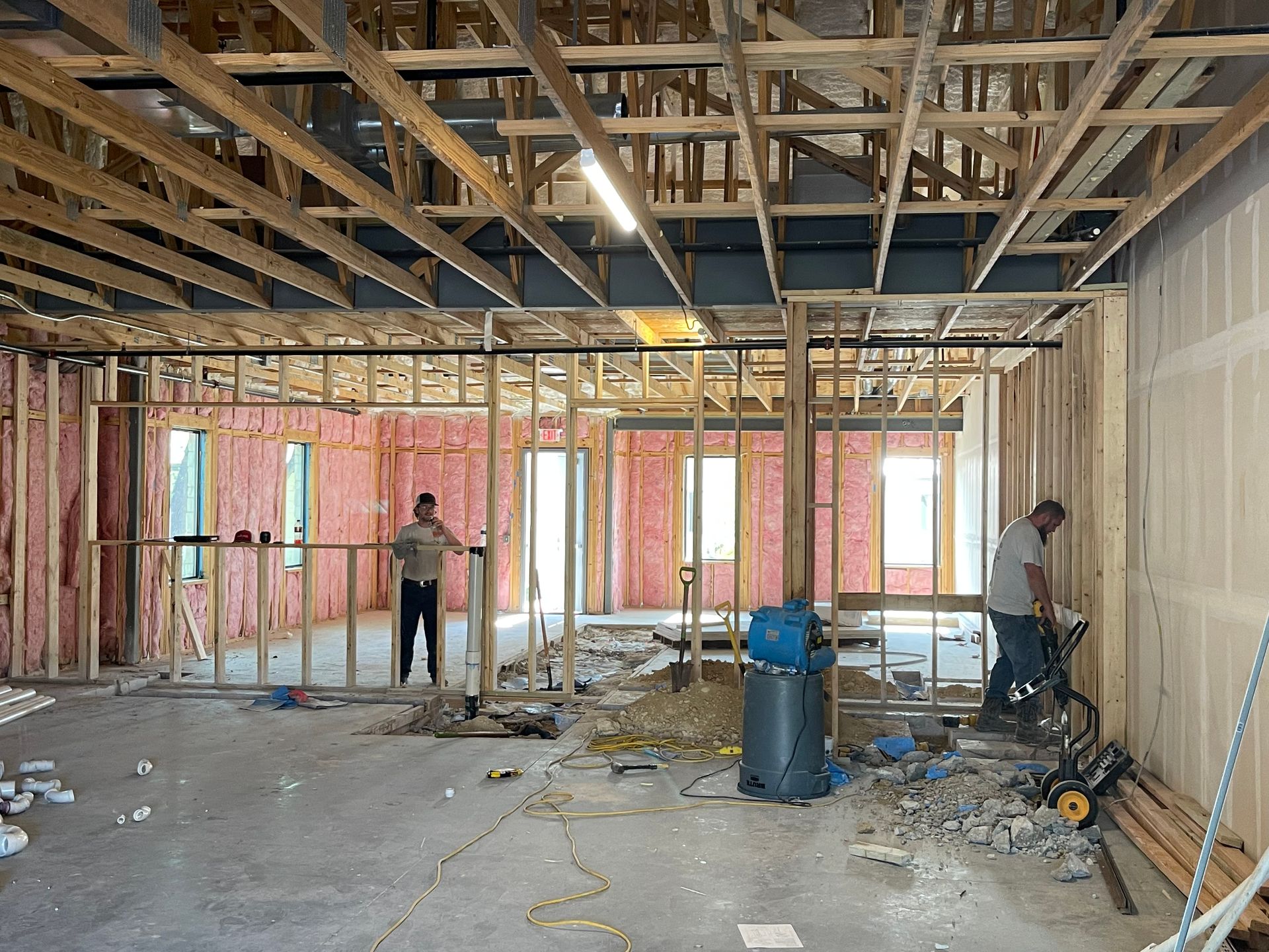 Partially framed interior under construction with exposed ceiling joists and two workers among tools and debris