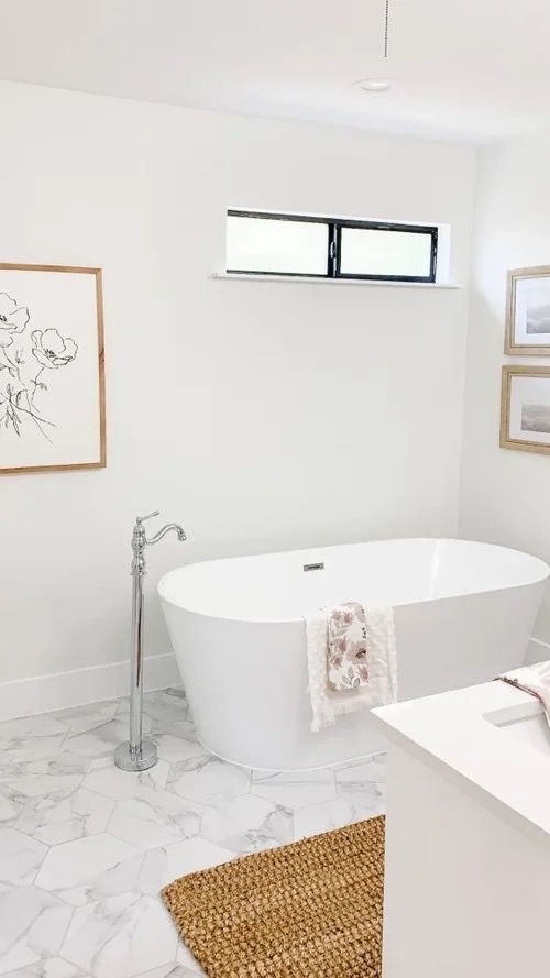 Bright white bathroom with a freestanding tub, marble floor, small window, and woven rug.