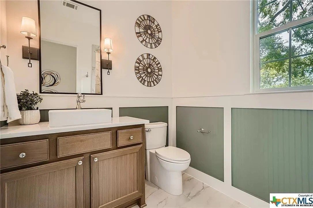 Elegant bathroom with wood vanity, white toilet, green paneling, and large window with trees outside