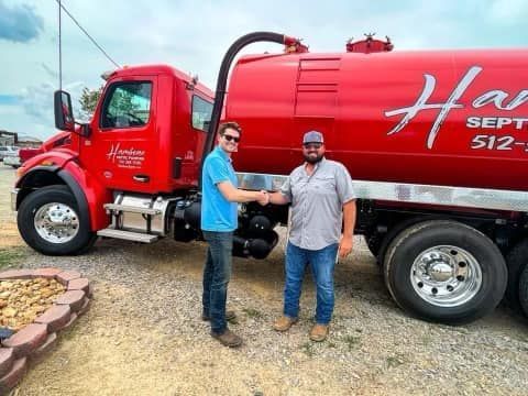 Two men shaking hands beside a red septic truck outdoors