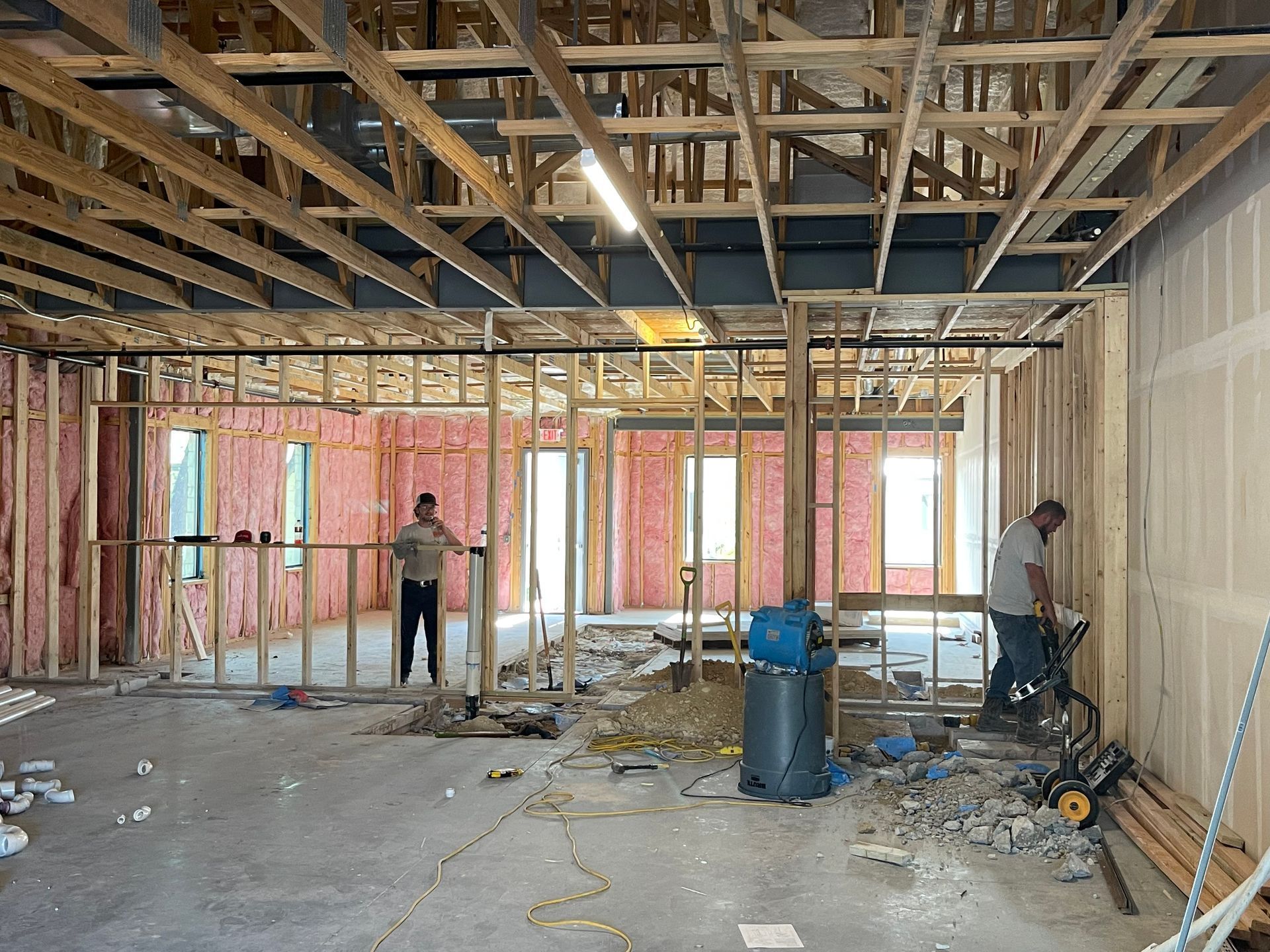 Unfinished room under construction with exposed framing, two workers, and scattered tools and debris