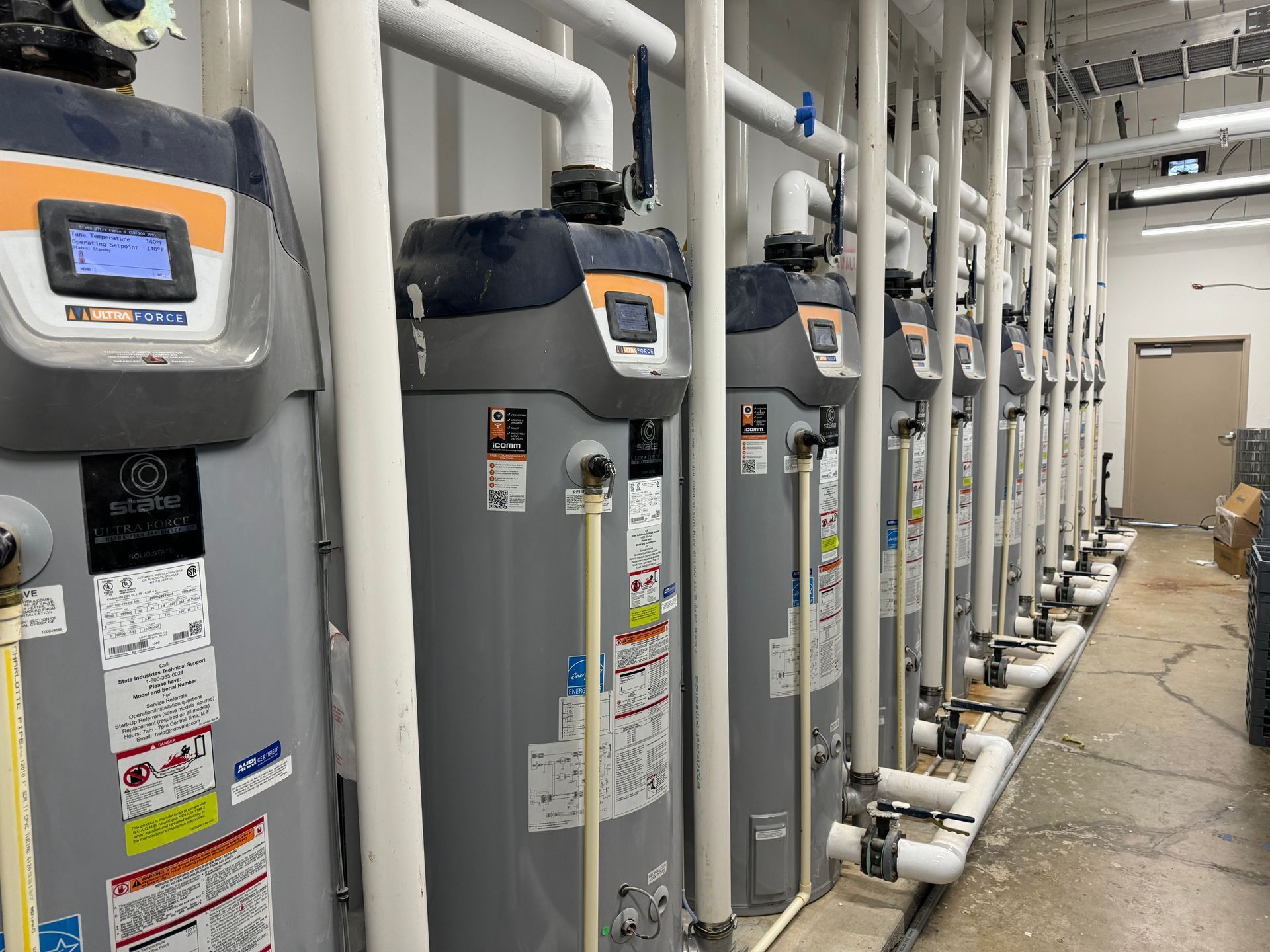 Row of gray water heaters and pipes in a mechanical room with a concrete floor and doorway at the back