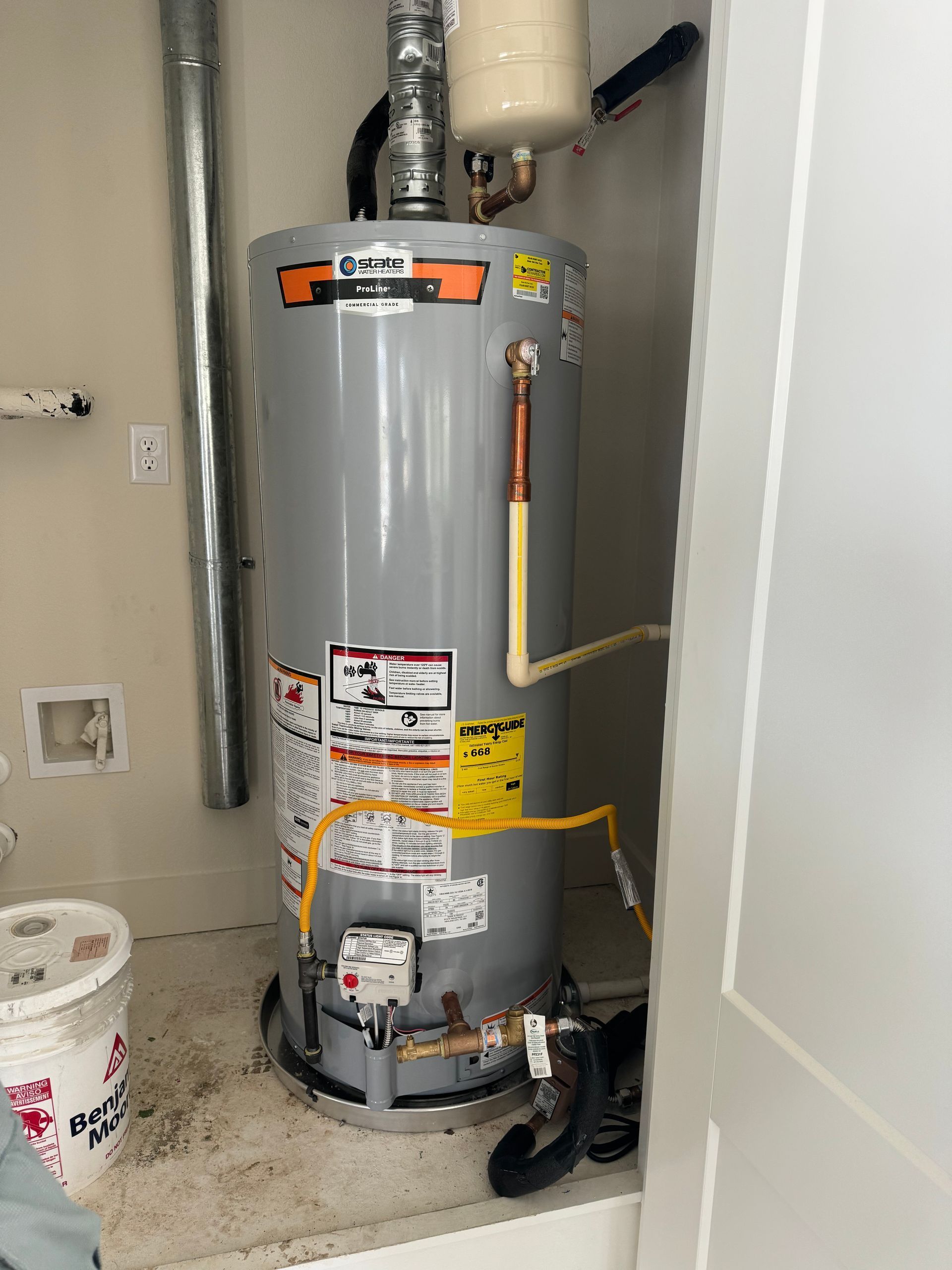 Gray water heater in a utility closet with exposed pipes and warning labels.