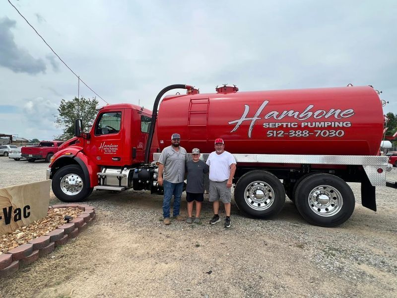 Two men standing beside a red vacuum tanker truck labeled “Hamp…” on a gravel lot
