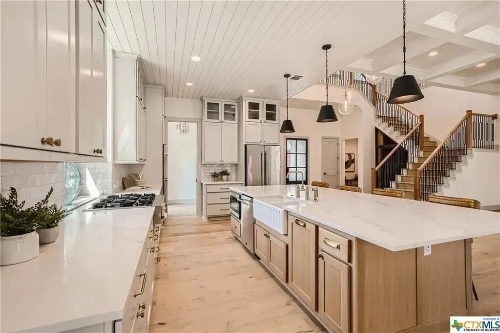 Bright white kitchen with large island, wood floors, black pendant lights, and staircase in the background