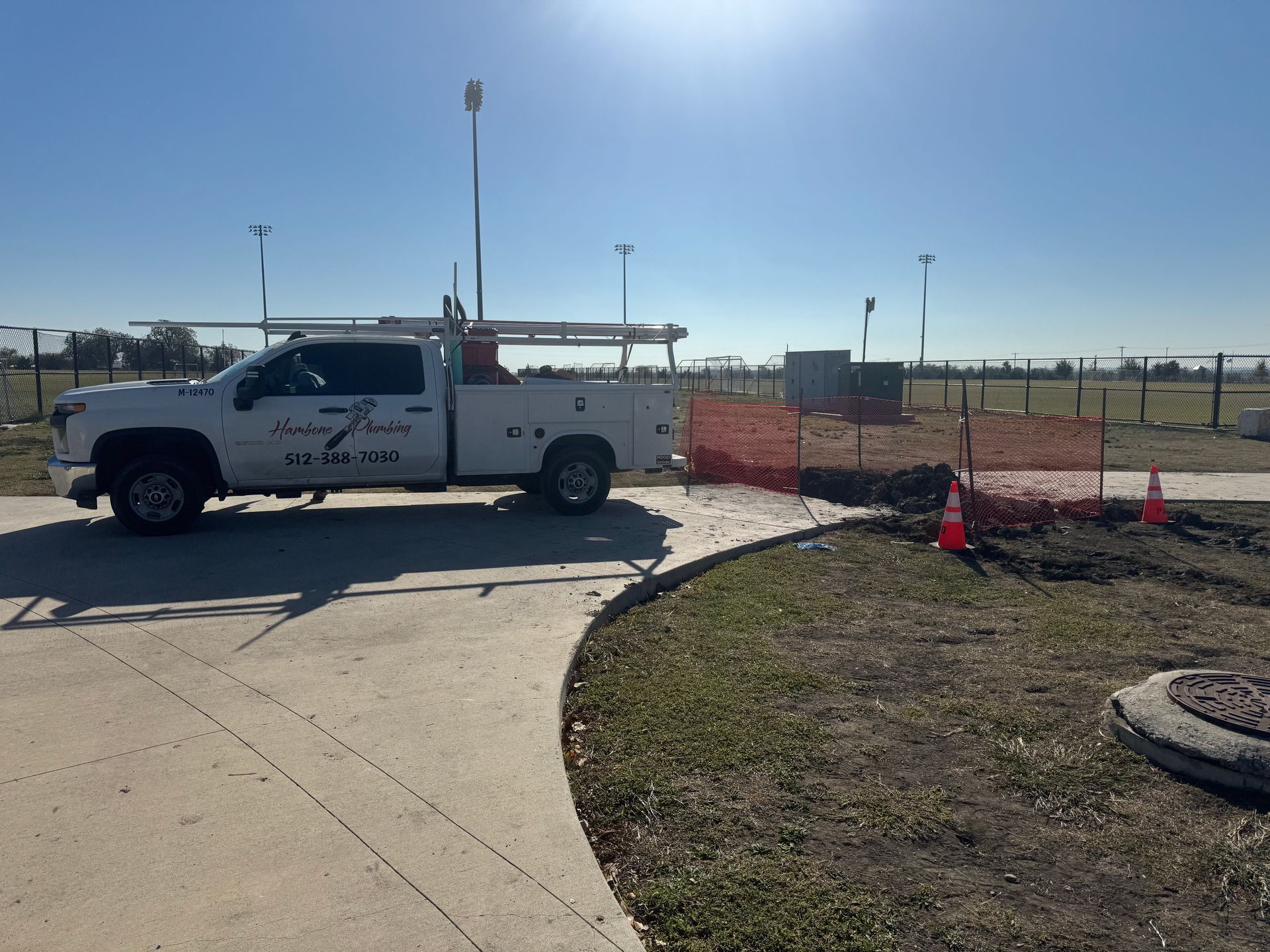White utility truck parked by a fenced roadside work area with orange cones and a dirt patch