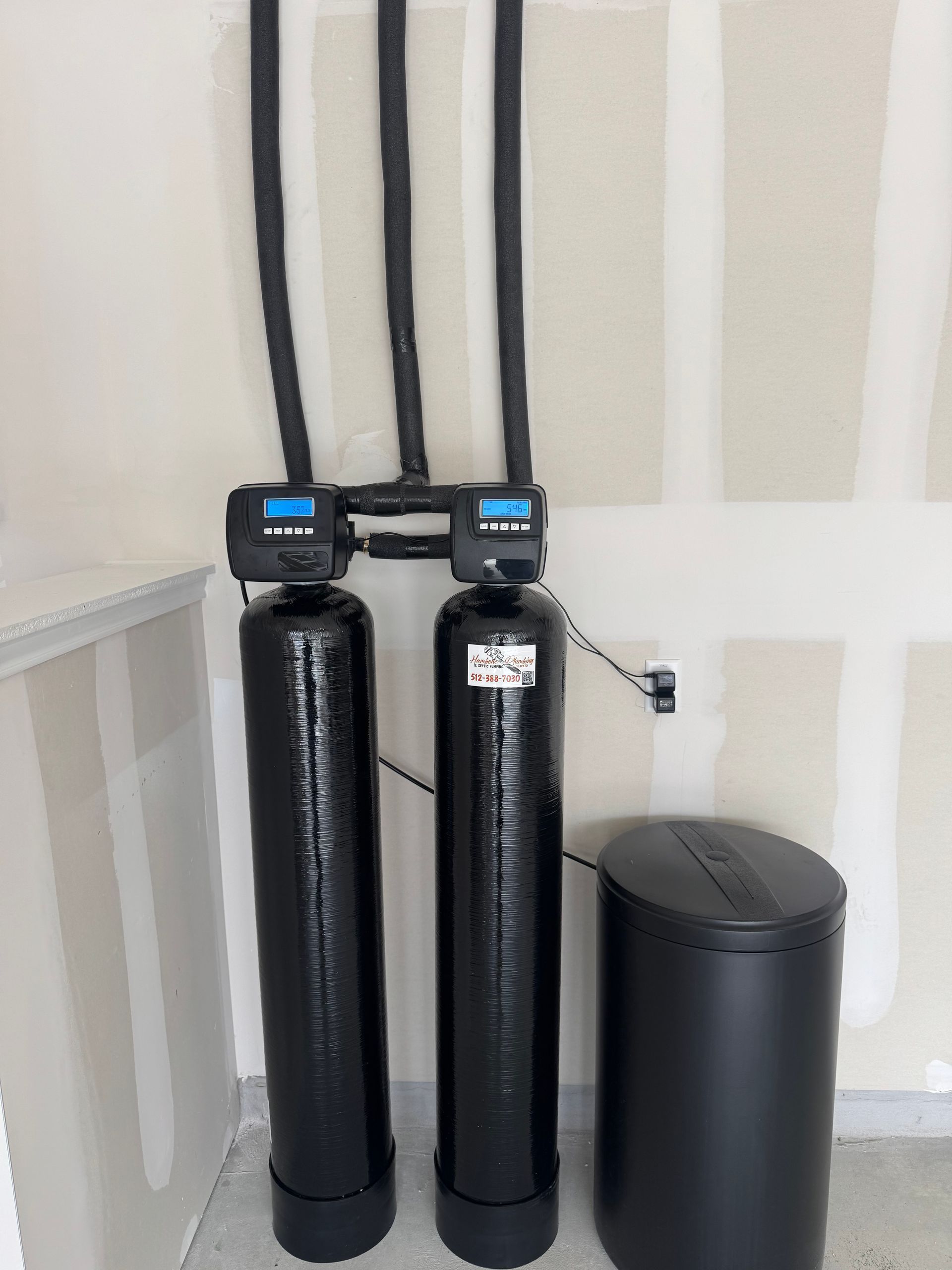 Two black water softener tanks and a black storage bin in a corner with beige walls and exposed pipes.