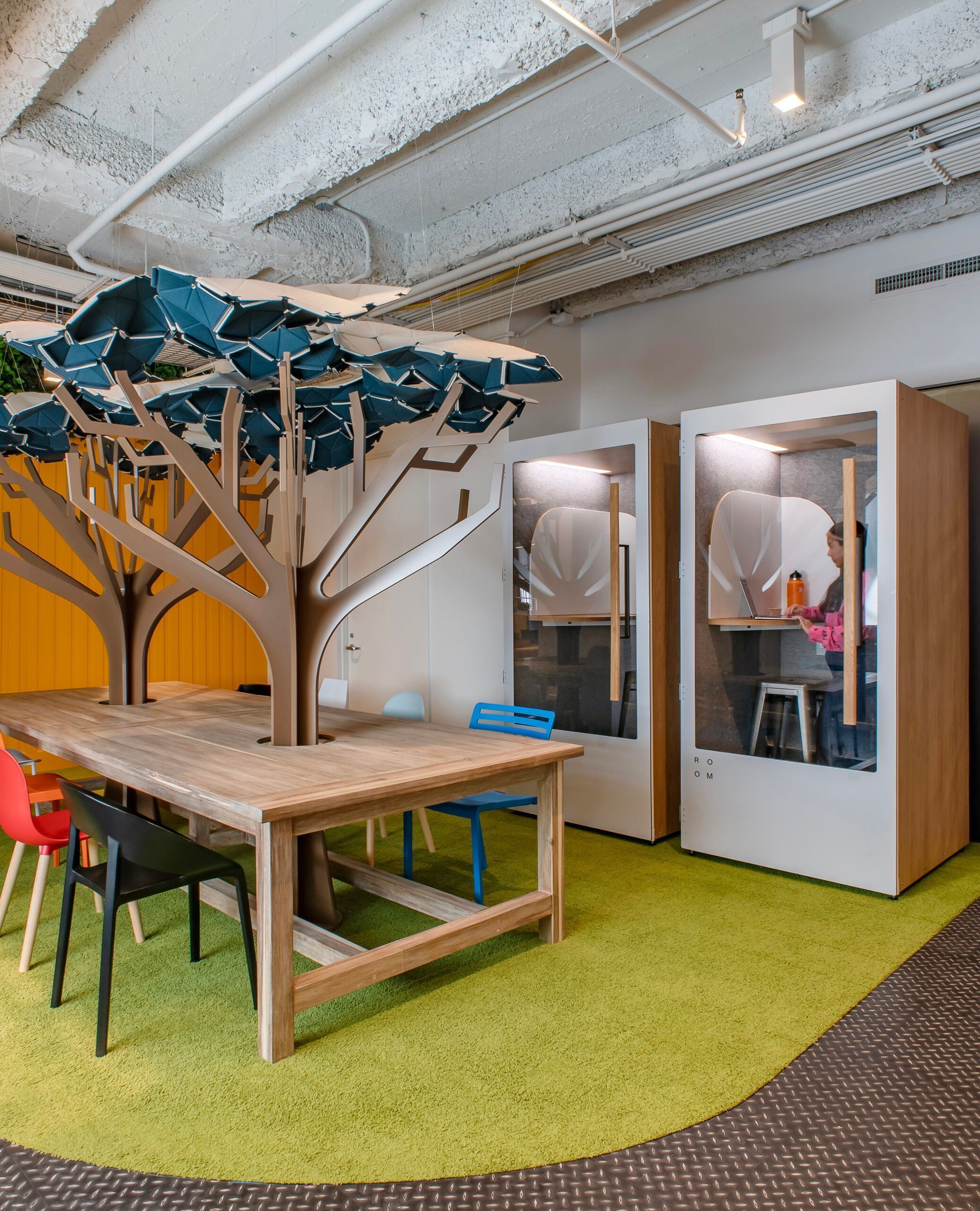 Office area with a wooden table, tree-shaped design, and soundproof booths; green carpet, yellow wall.