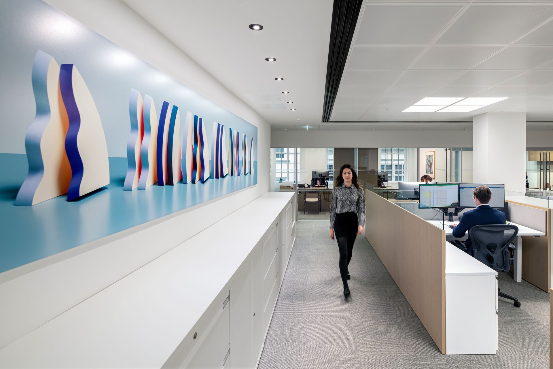 Woman walking in modern office, past a long counter with artwork and cubicles with workers.