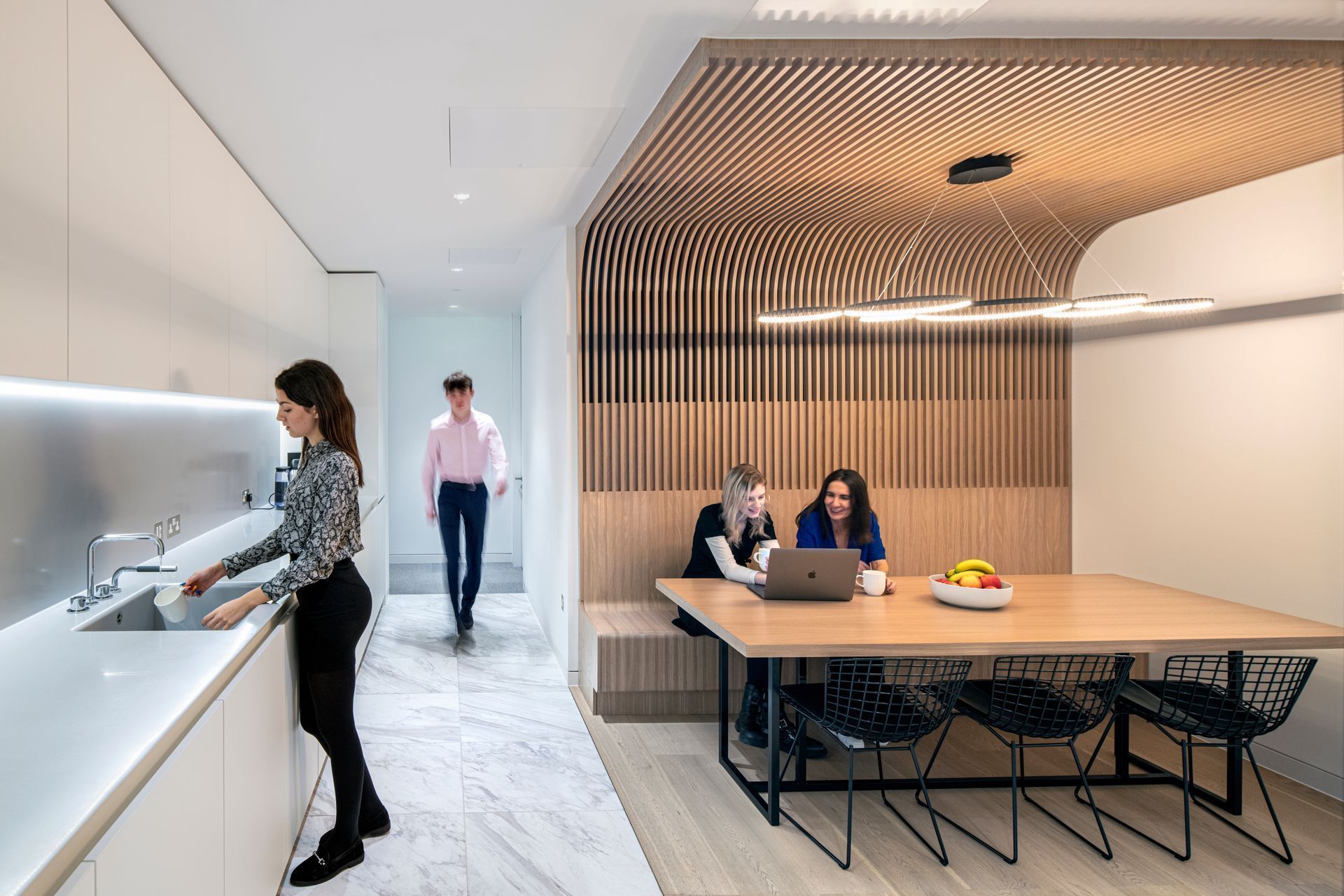 Modern office interior with people: a woman at a sink, two at a table, and one walking. Light wood and white.