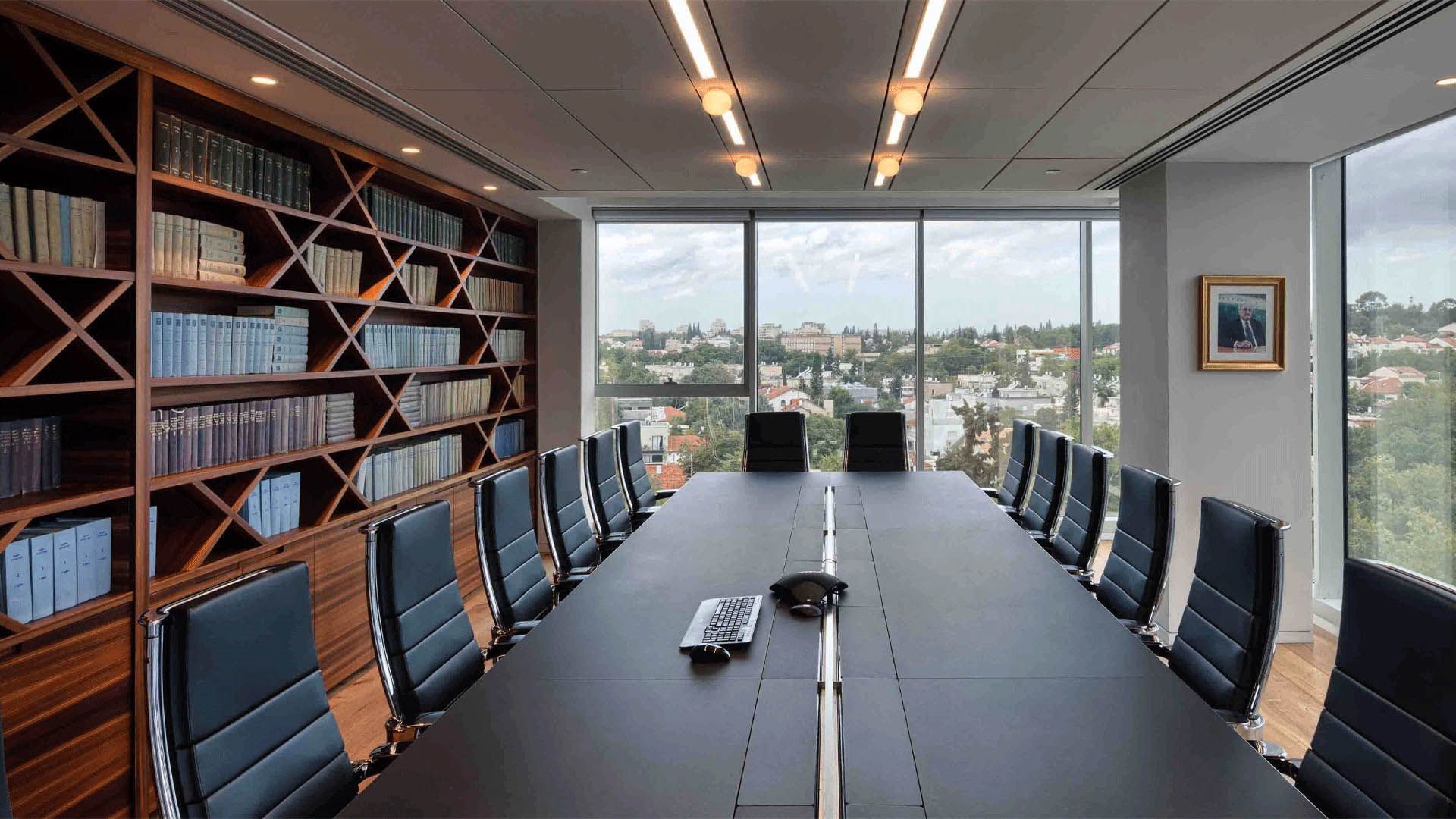 Conference room with long black table, black chairs, bookcase, and cityscape view.