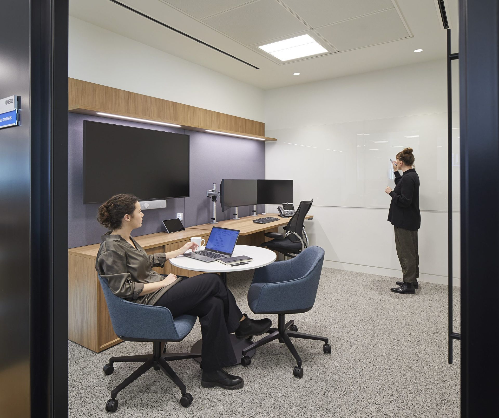 Two people in a modern office room: one seated at a table with a laptop, the other standing at a whiteboard, pointing.