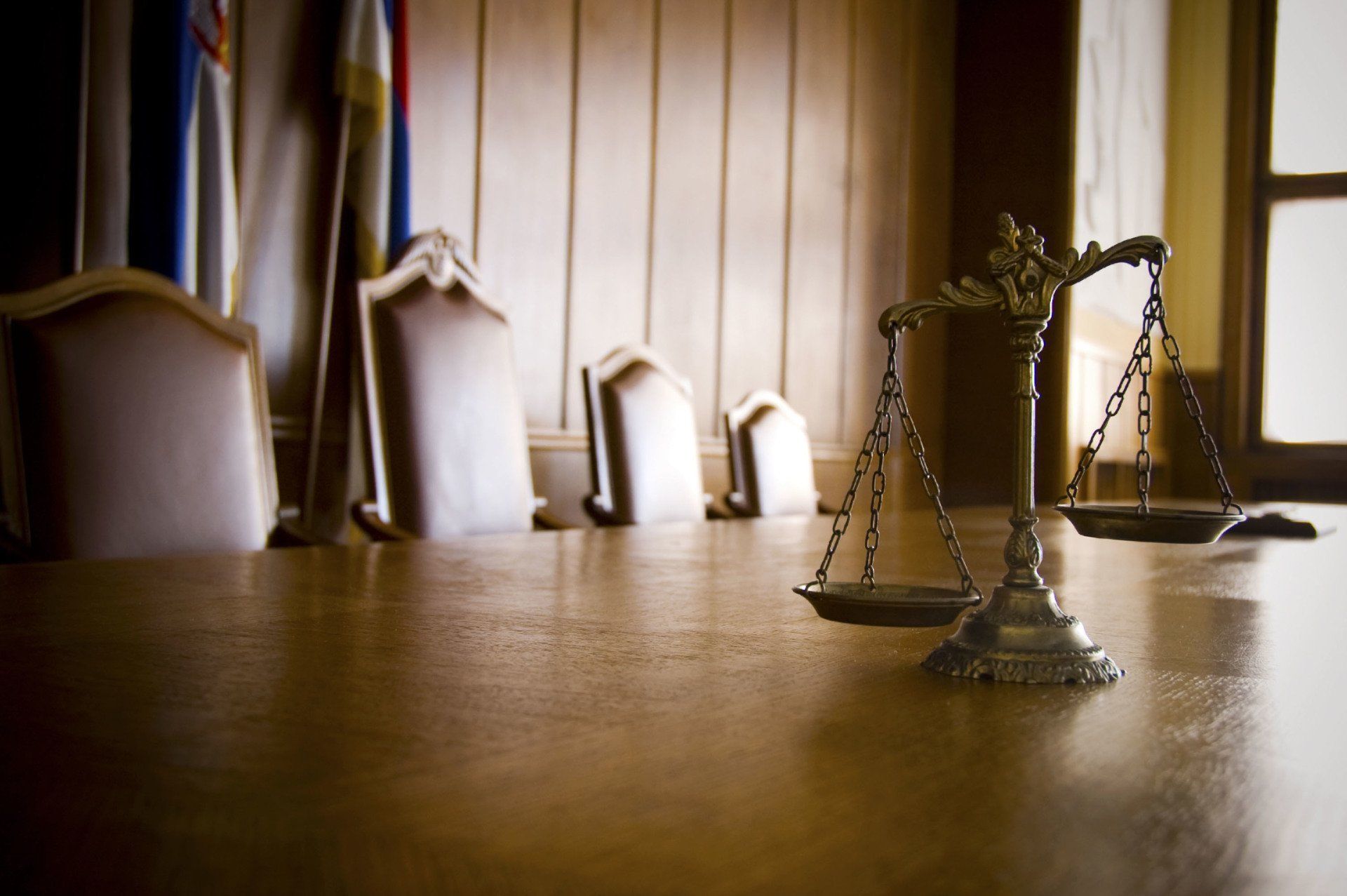 Justice scales on a wooden table in a courtroom, with empty chairs and flags in the background.