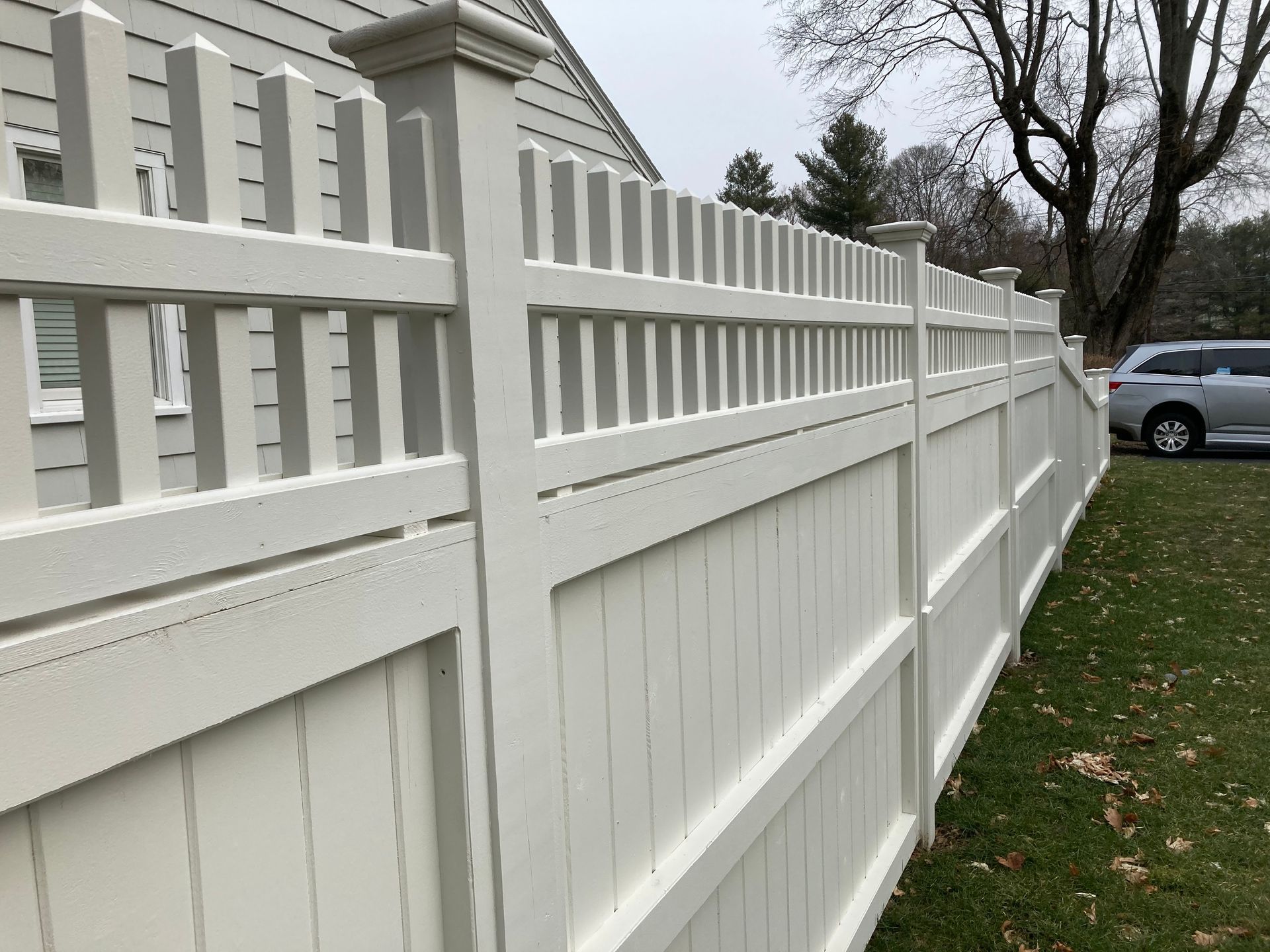 A white fence is surrounding a house with a car parked behind it.
