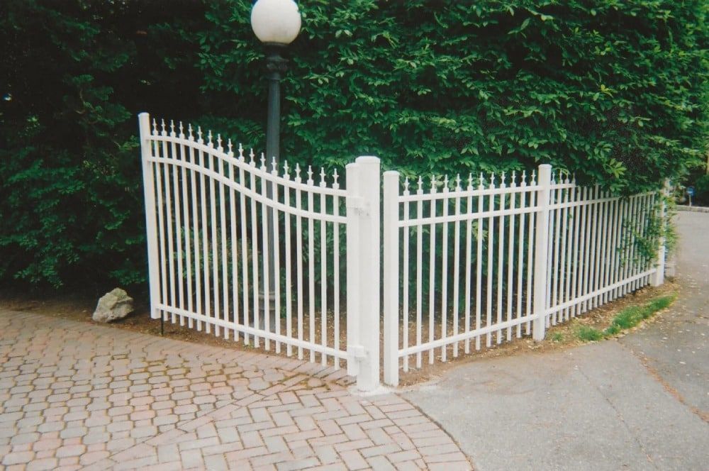A white fence surrounds a brick driveway with trees in the background