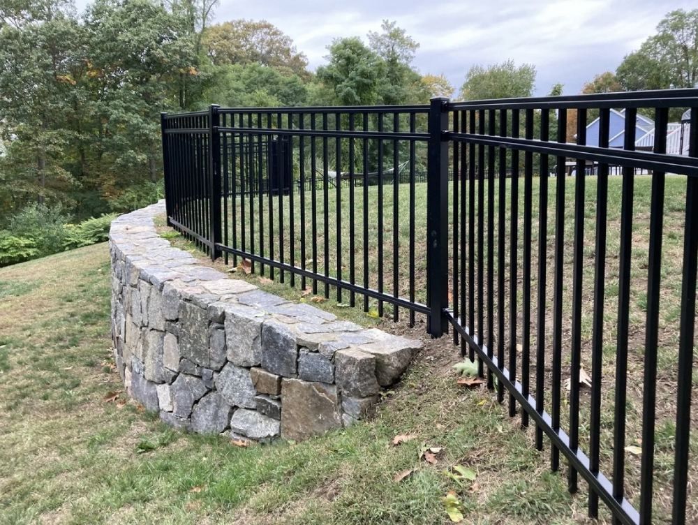A black metal fence surrounds a stone wall in a yard.
