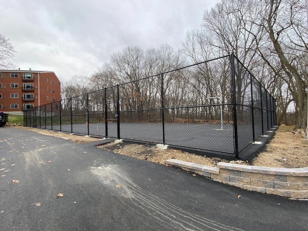A chain link fence surrounds a basketball court in a park.