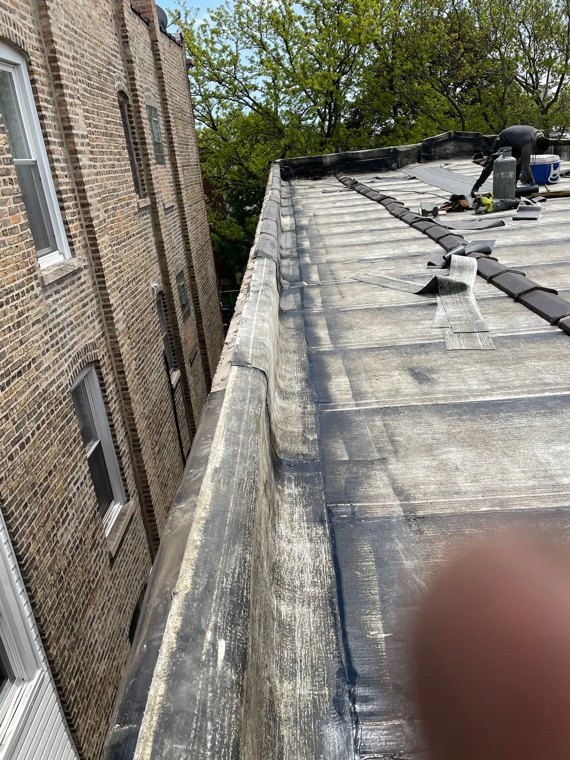 Rooftop with black tar surface next to a brick building. Trees in background.