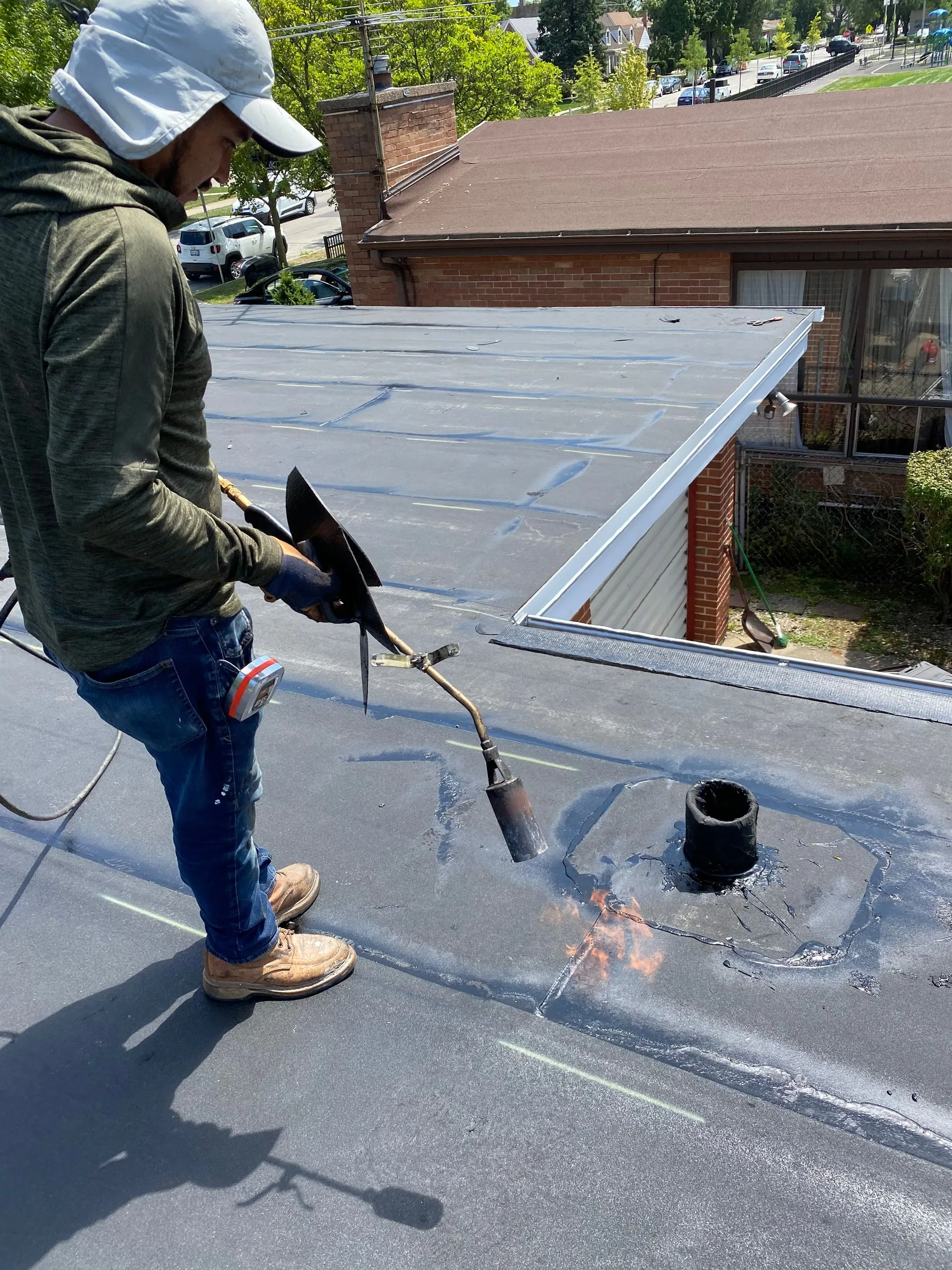 Person using a torch on a flat roof. They wear work clothes and a hat. Rooftop setting, daylight.